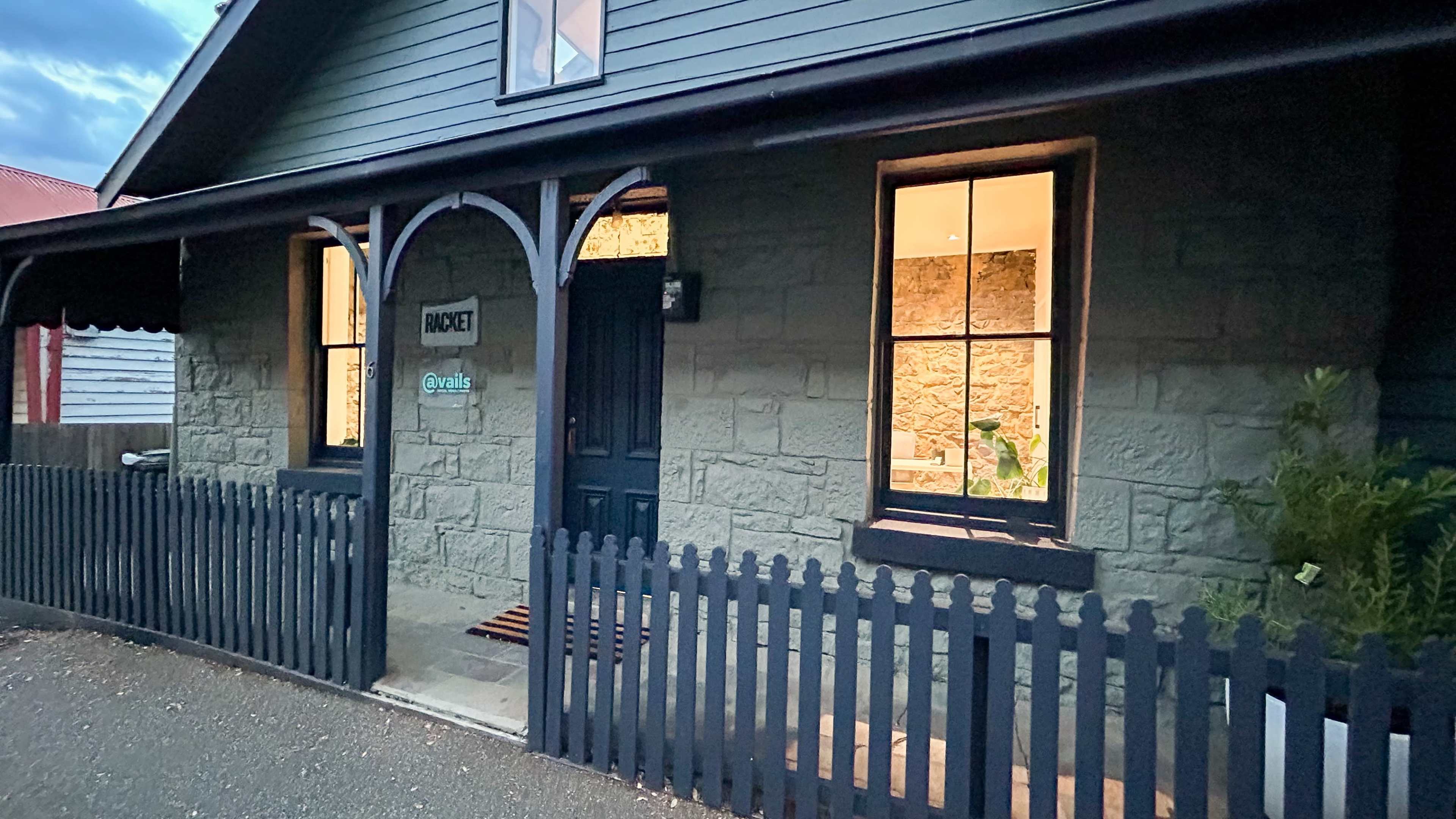 A stone house with a black door and window frames, featuring a wooden fence and a sign that reads "INQUIRY" on the exterior.