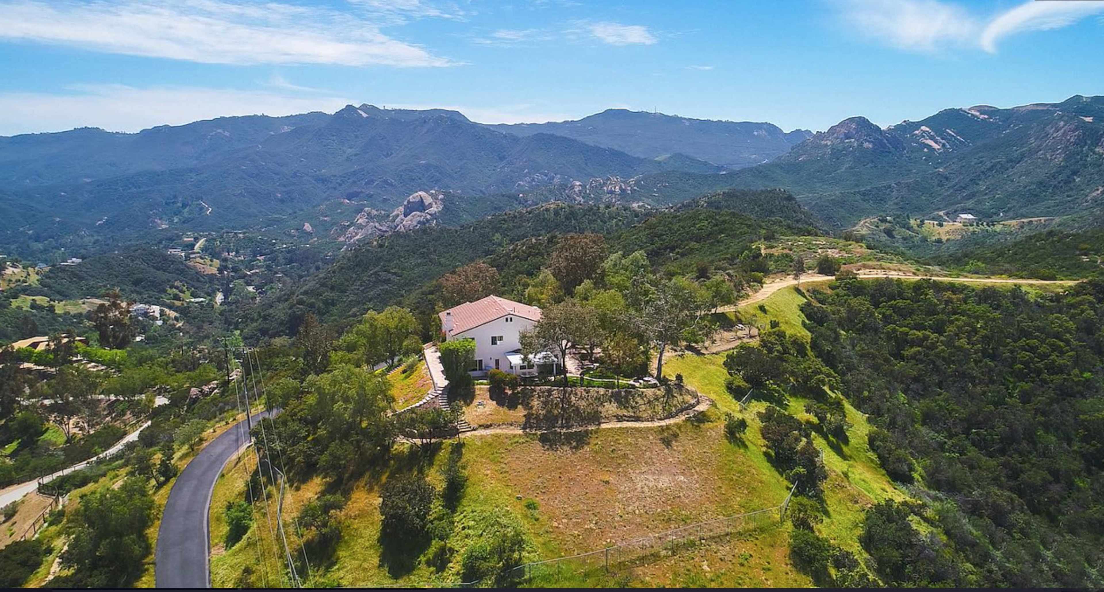 A single-story house is situated on a hillside, surrounded by trees and overlooking rolling mountains and valleys under a clear blue sky.