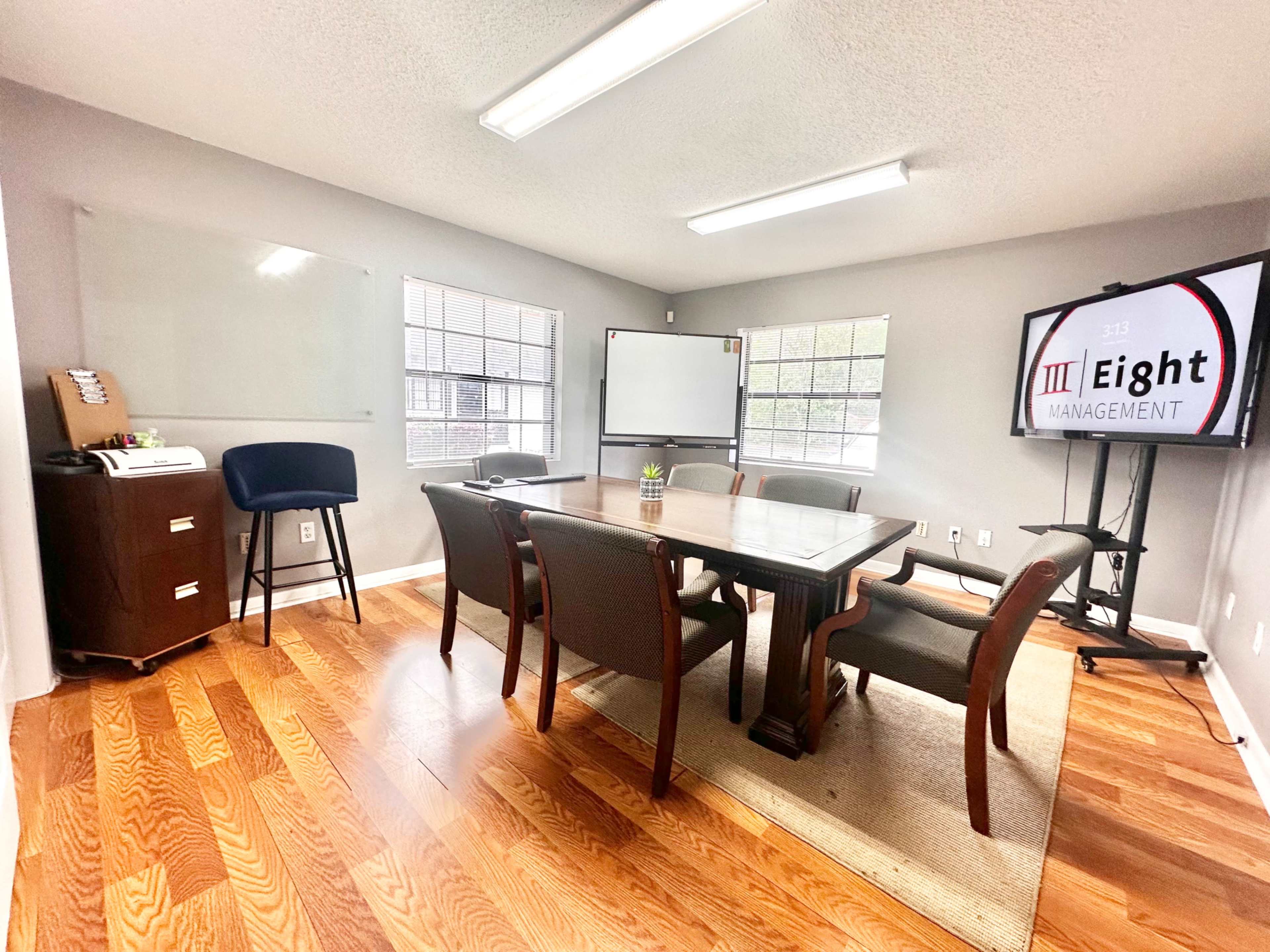 A conference room features a rectangular table surrounded by chairs, a large screen displaying "Eight Management," and windows allowing natural light.