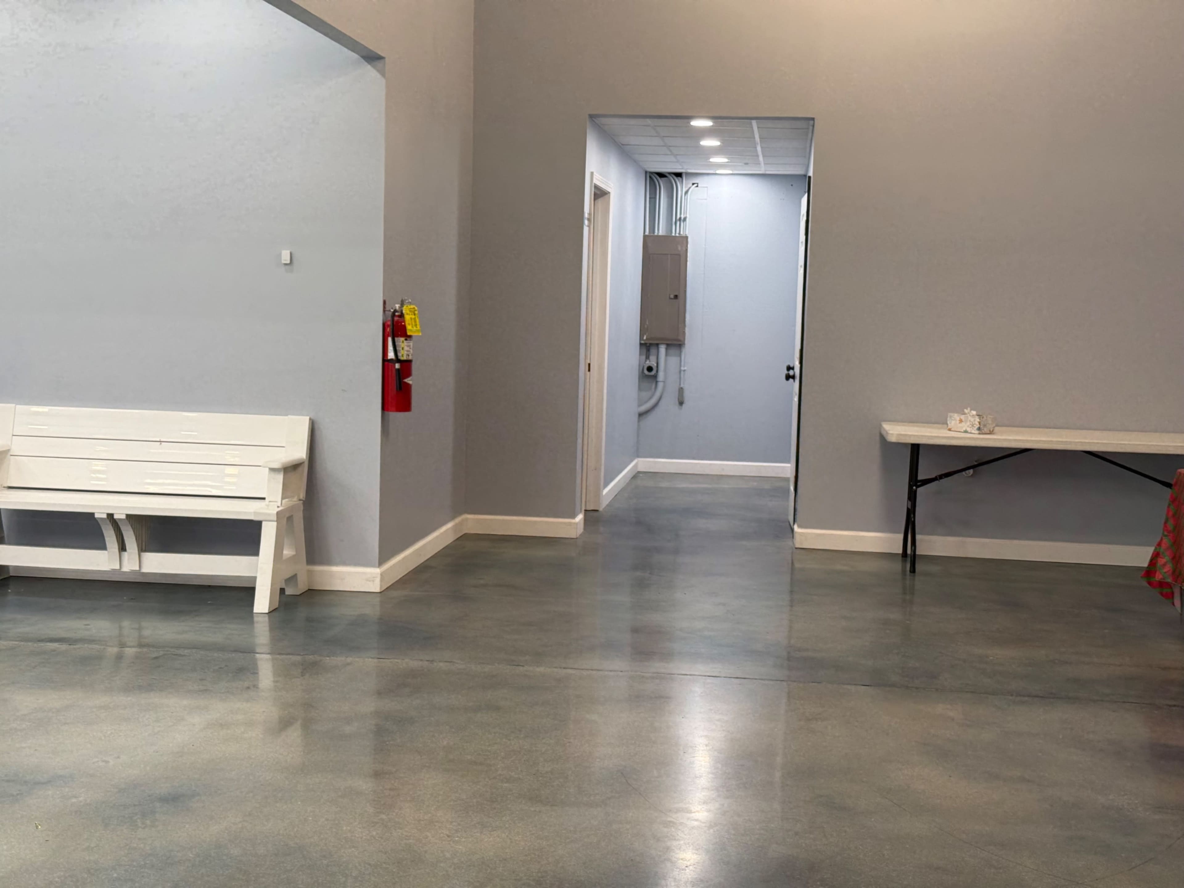 The image shows a spacious, well-lit interior with a gray wall, a white bench, and a corridor leading to a door at the end.