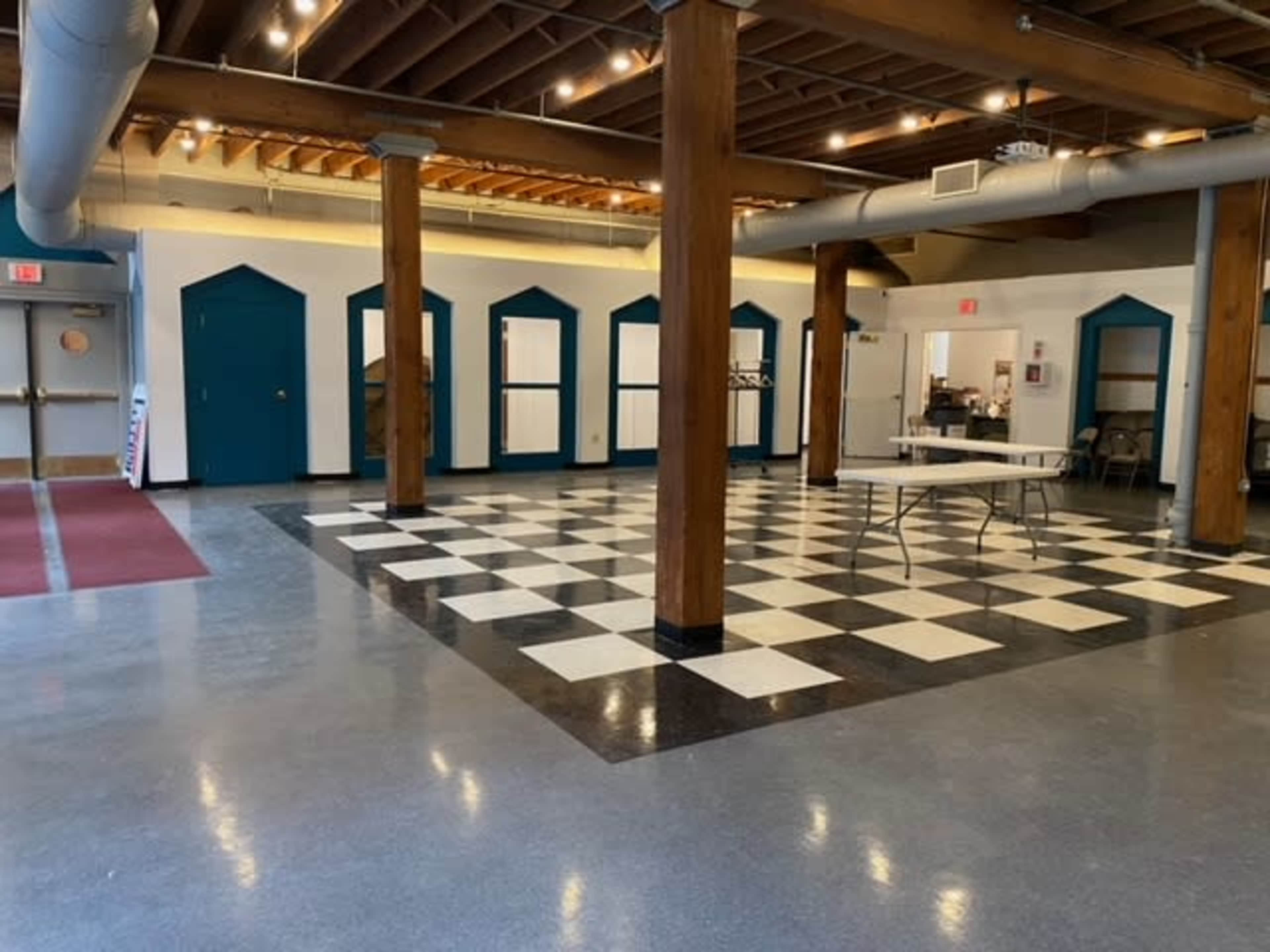 The image shows a spacious indoor area with a checkered floor, wooden pillars, and doorways leading to other rooms.