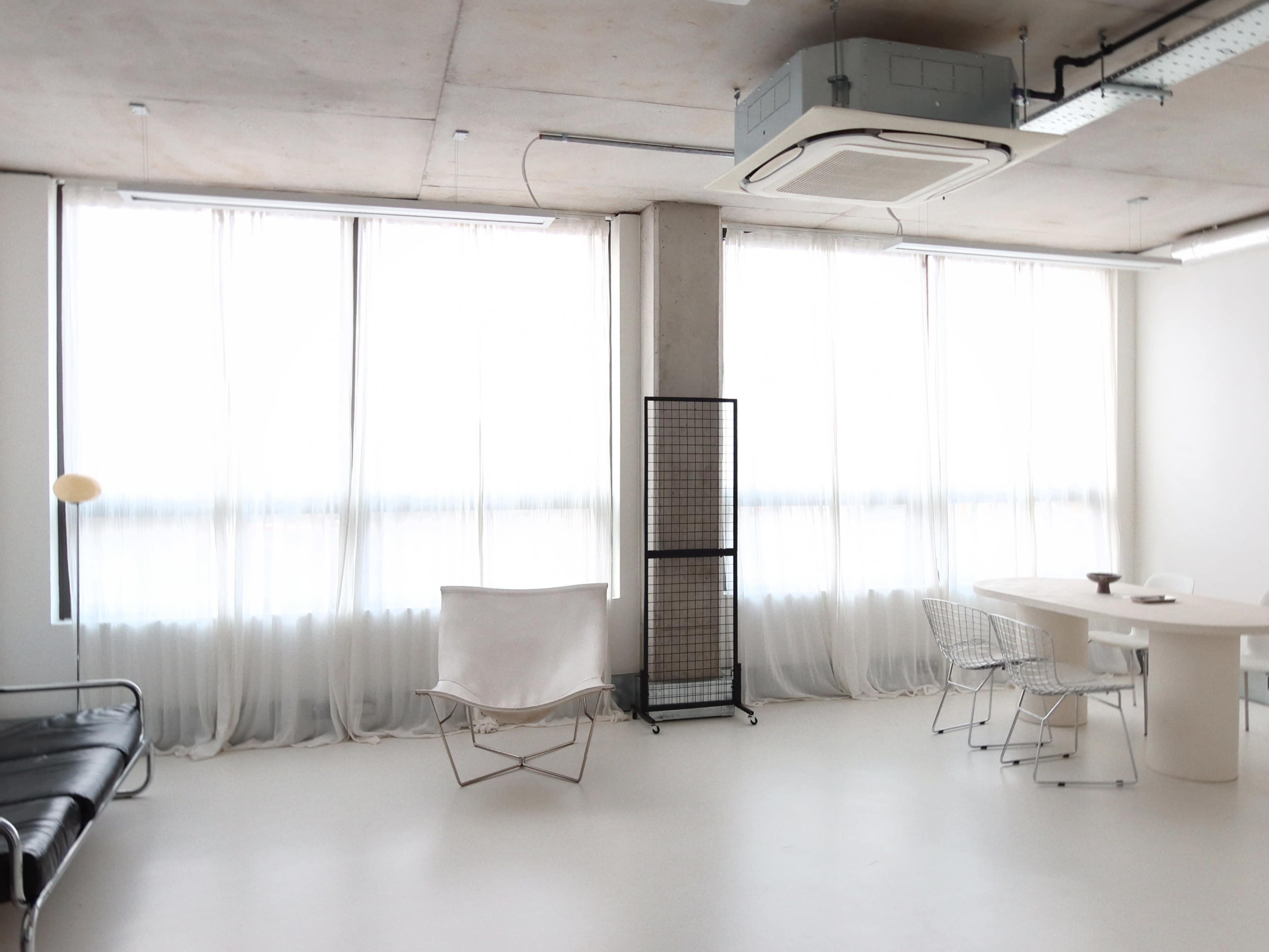 The image shows a minimalist room with large windows draped in sheer white curtains, featuring a black sofa, a white chair, a round dining table, and a metal folding screen.