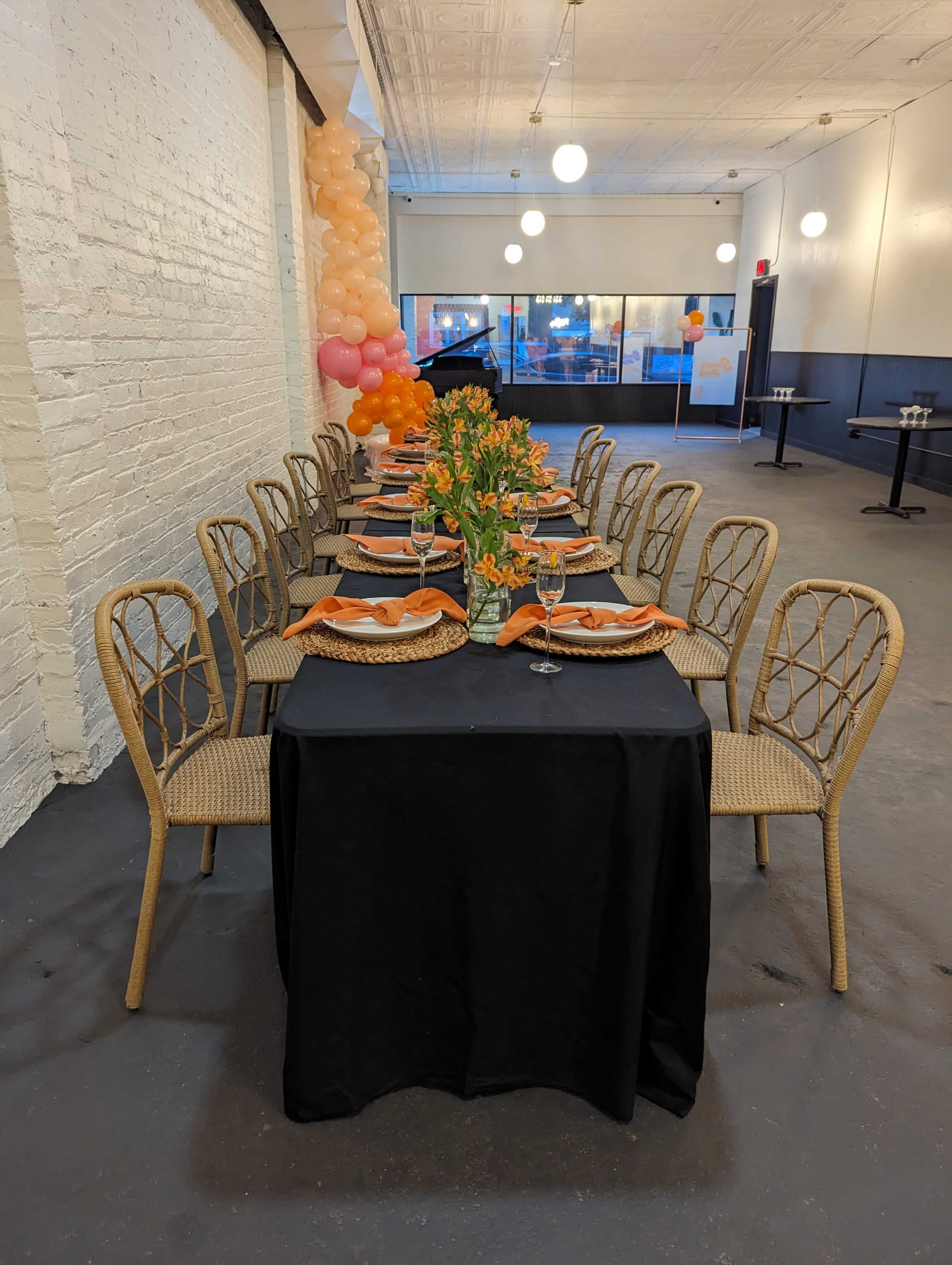 A long dining table set with orange and beige table settings, surrounded by rattan chairs, in a decorated space with balloon arrangements in the background.