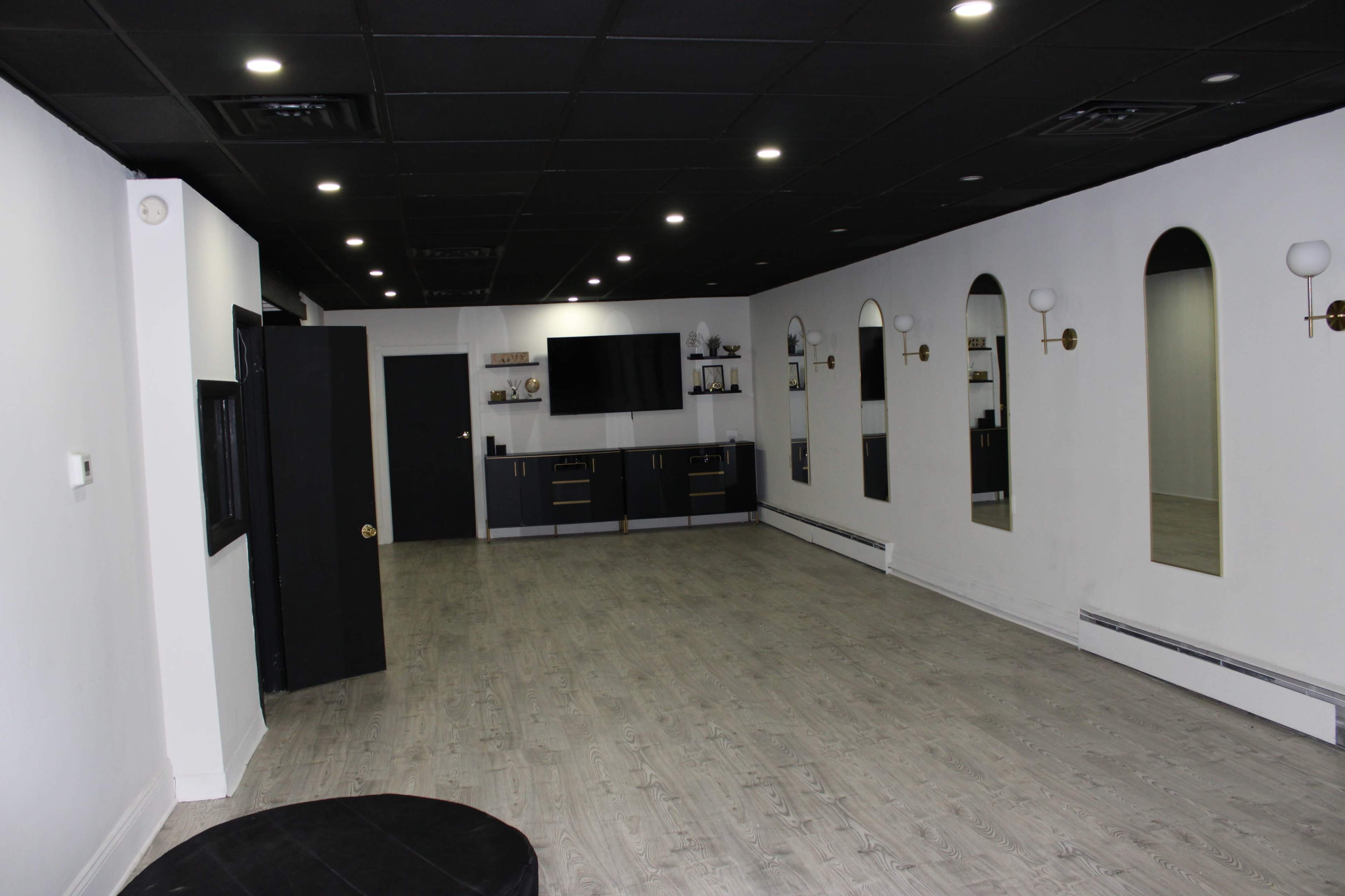 The image shows an empty, well-lit studio with light wood flooring, black accents, and several wall-mounted mirrors.