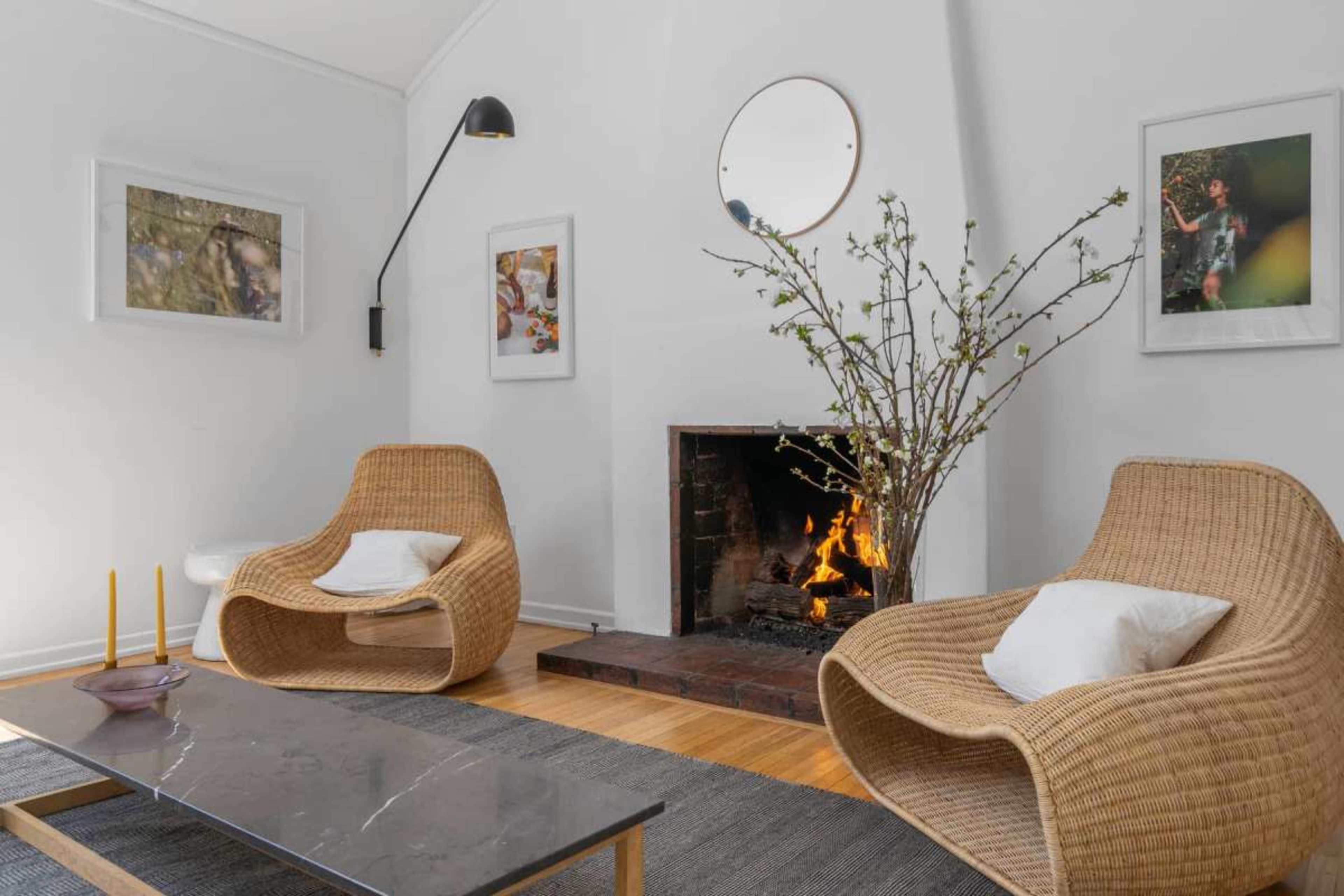 The image shows a cozy living room featuring two woven chairs, a coffee table, and a fireplace with a fire burning, decorated with a branch arrangement and framed photos on the walls.