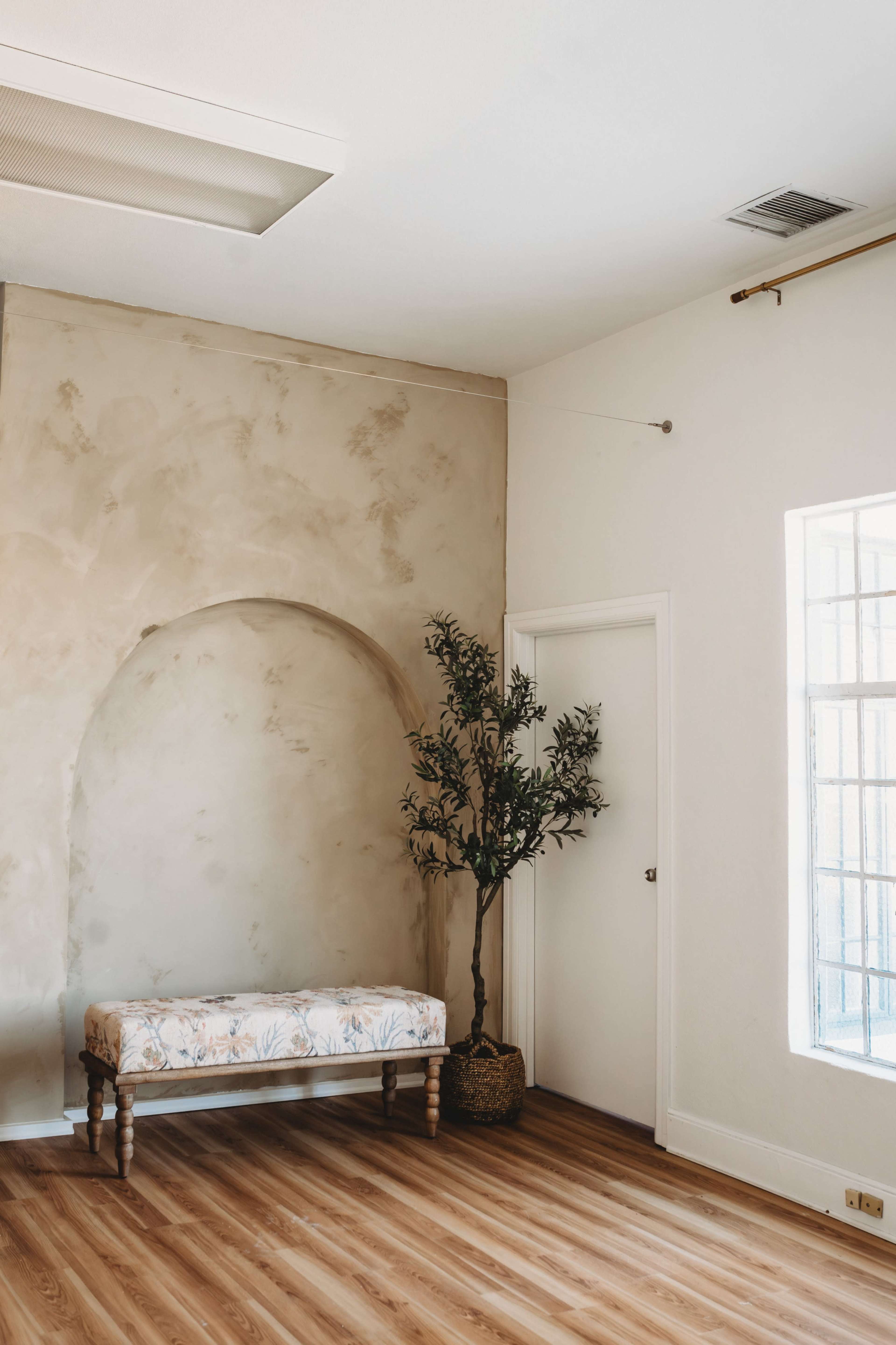 The image shows a minimalist room featuring a patterned bench, a decorative plant in a basket, and a wall with an arched recess.