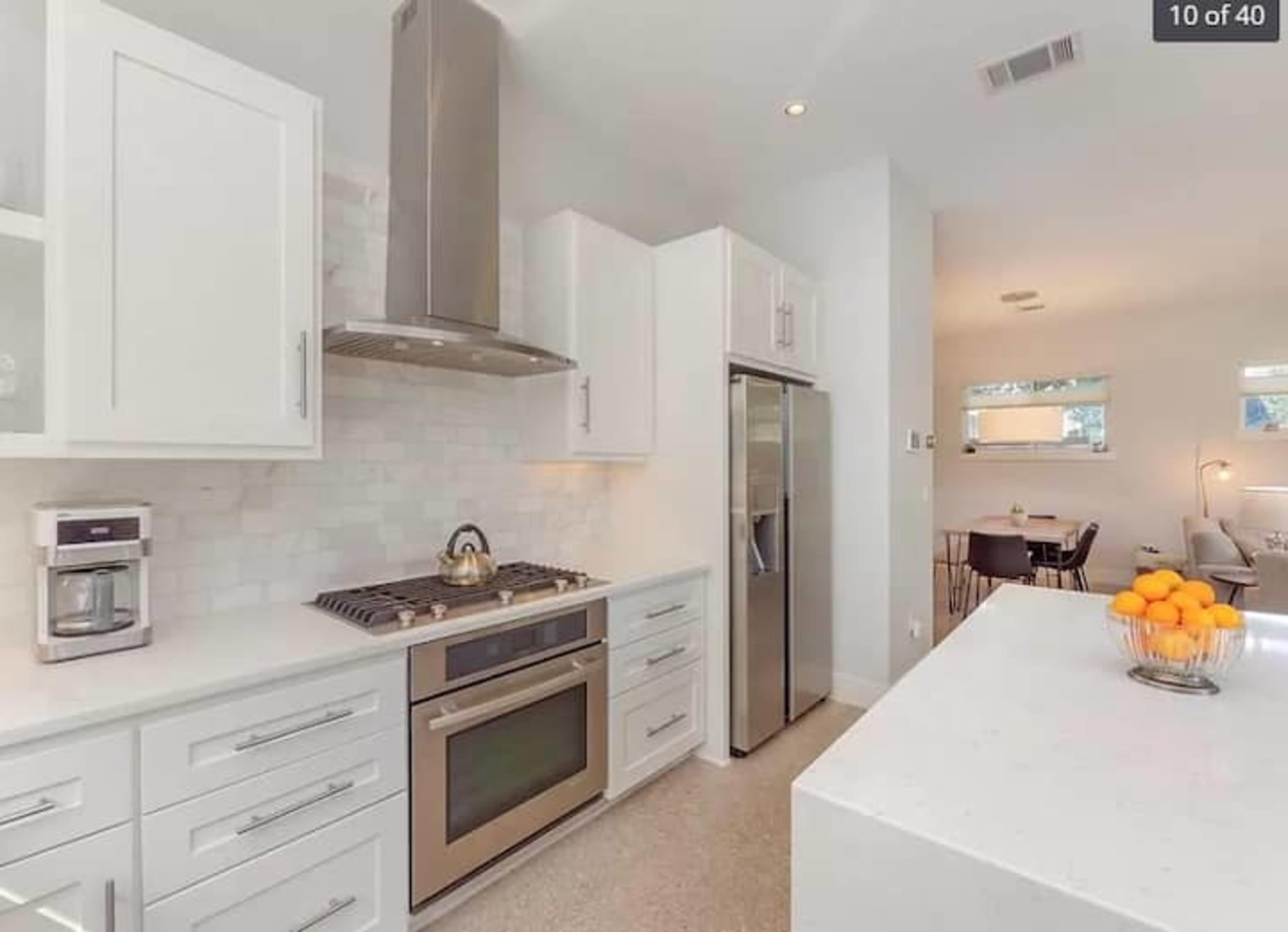 A modern kitchen features white cabinetry, stainless steel appliances, a gas stove, and a large island with a bowl of oranges.