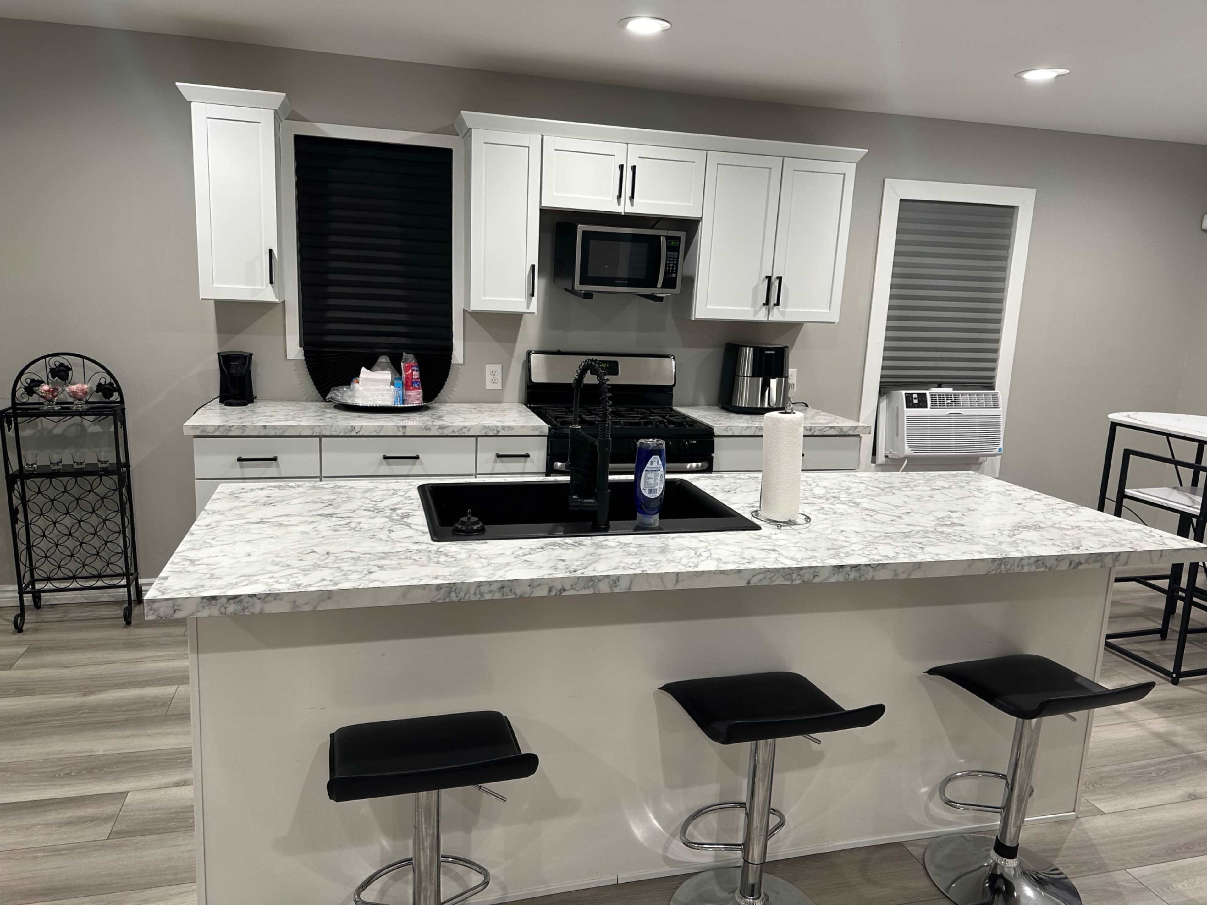 The image shows a modern kitchen with white cabinets, a marble countertop, a black sink, and four black bar stools.