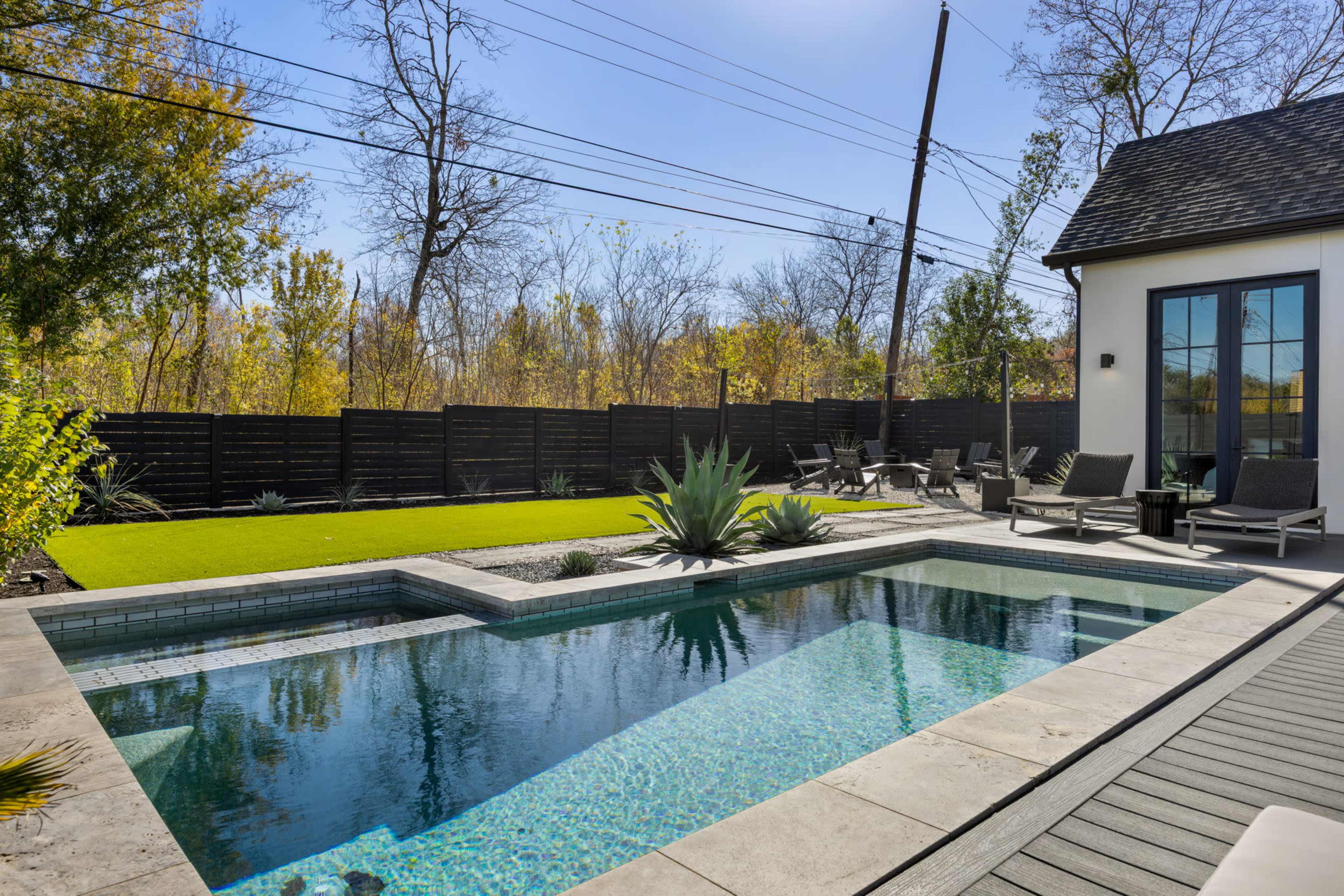 The image shows a modern backyard featuring a rectangular swimming pool, surrounded by a stone deck and lounge chairs, with greenery and trees in the background.