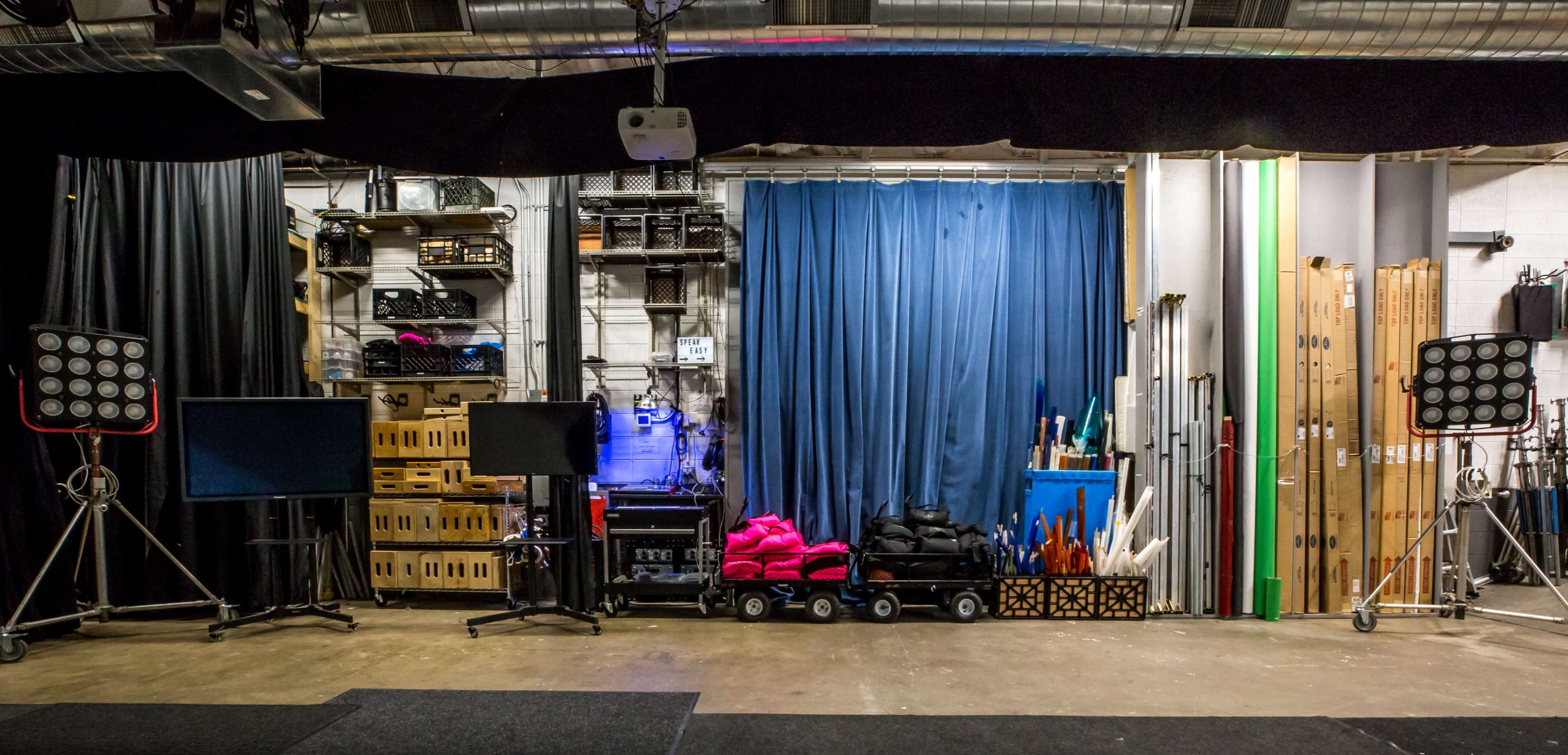 A production studio with a blue curtain backdrop, storage bins, equipment shelves, and two lighting setups on tripods.