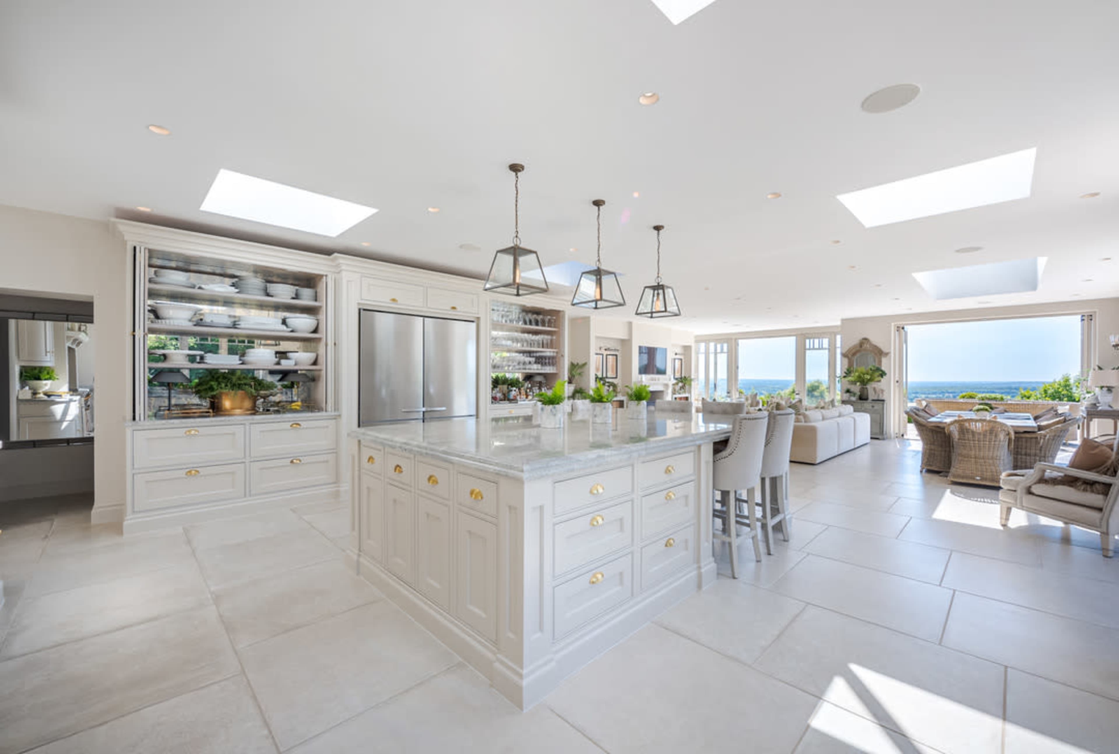 The image shows a bright, modern kitchen featuring a large island with a marble countertop, ample cabinetry, and views of the ocean from large windows.