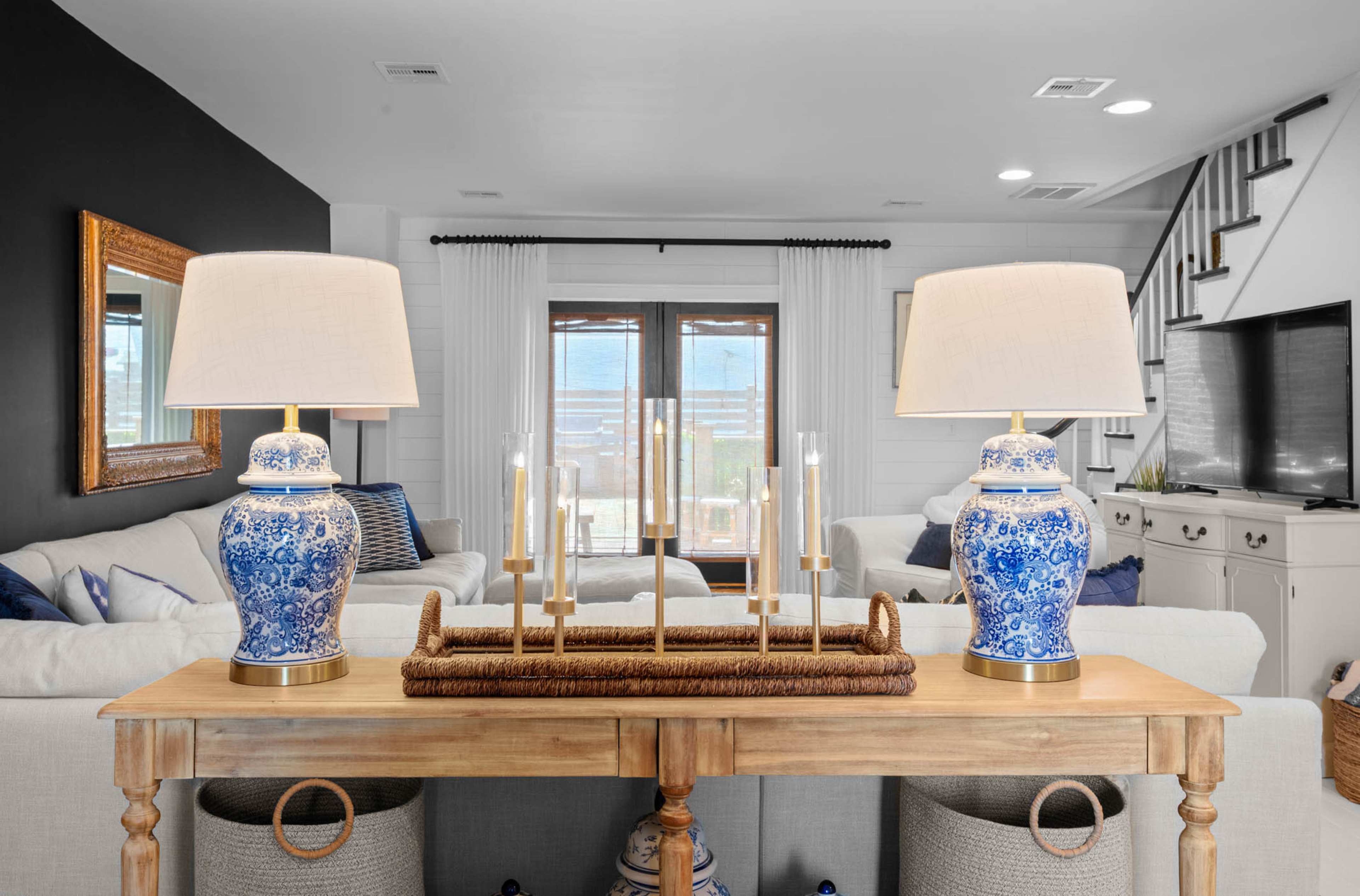 A wooden table with blue and white ceramic lamps and a decorative candle holder is placed in the foreground, while a living room with a sofa, TV, and patio doors is visible in the background.