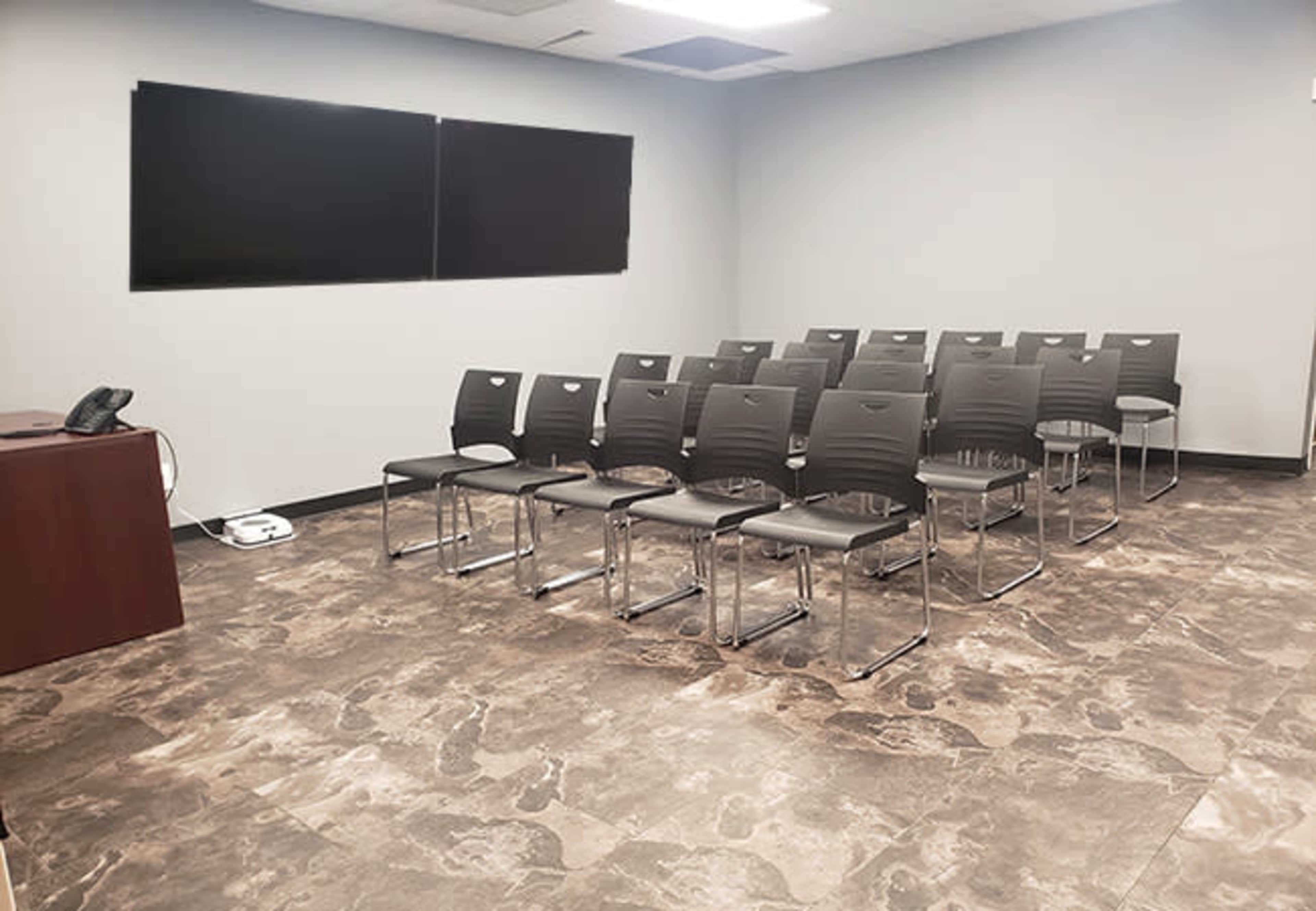 A conference room with a row of black chairs arranged in front of two large screens and a wooden table with a phone.