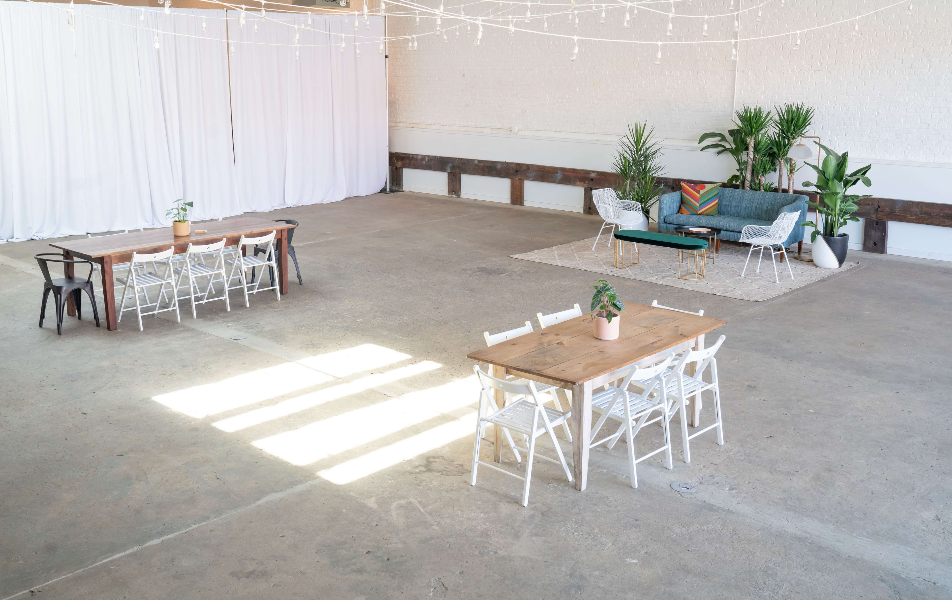 A spacious room with two dining tables, white chairs, a lounge area with a green couch, and decorative plants under string lights.