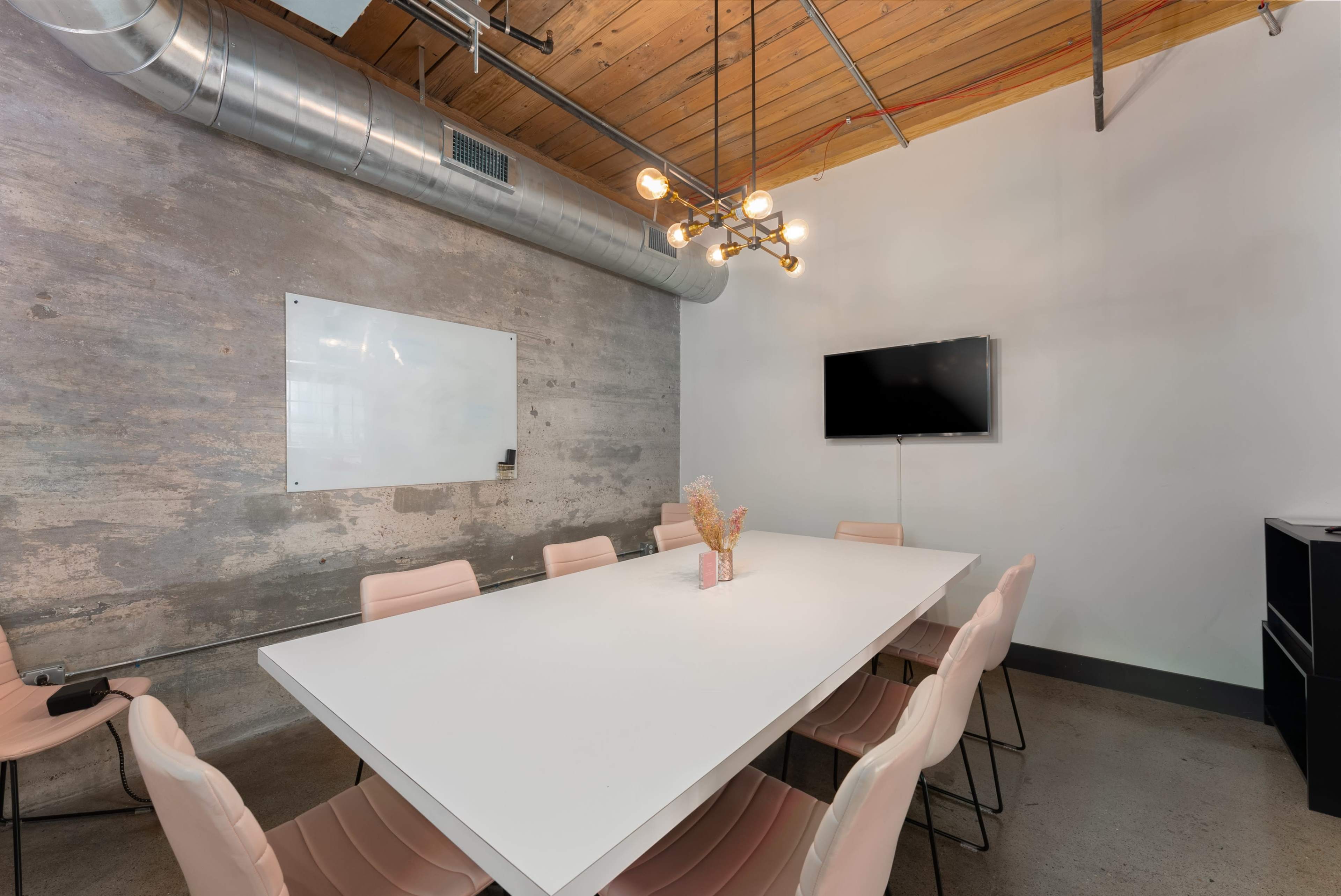 A modern conference room features a large white table surrounded by pink chairs, with a whiteboard and a TV mounted on the wall.