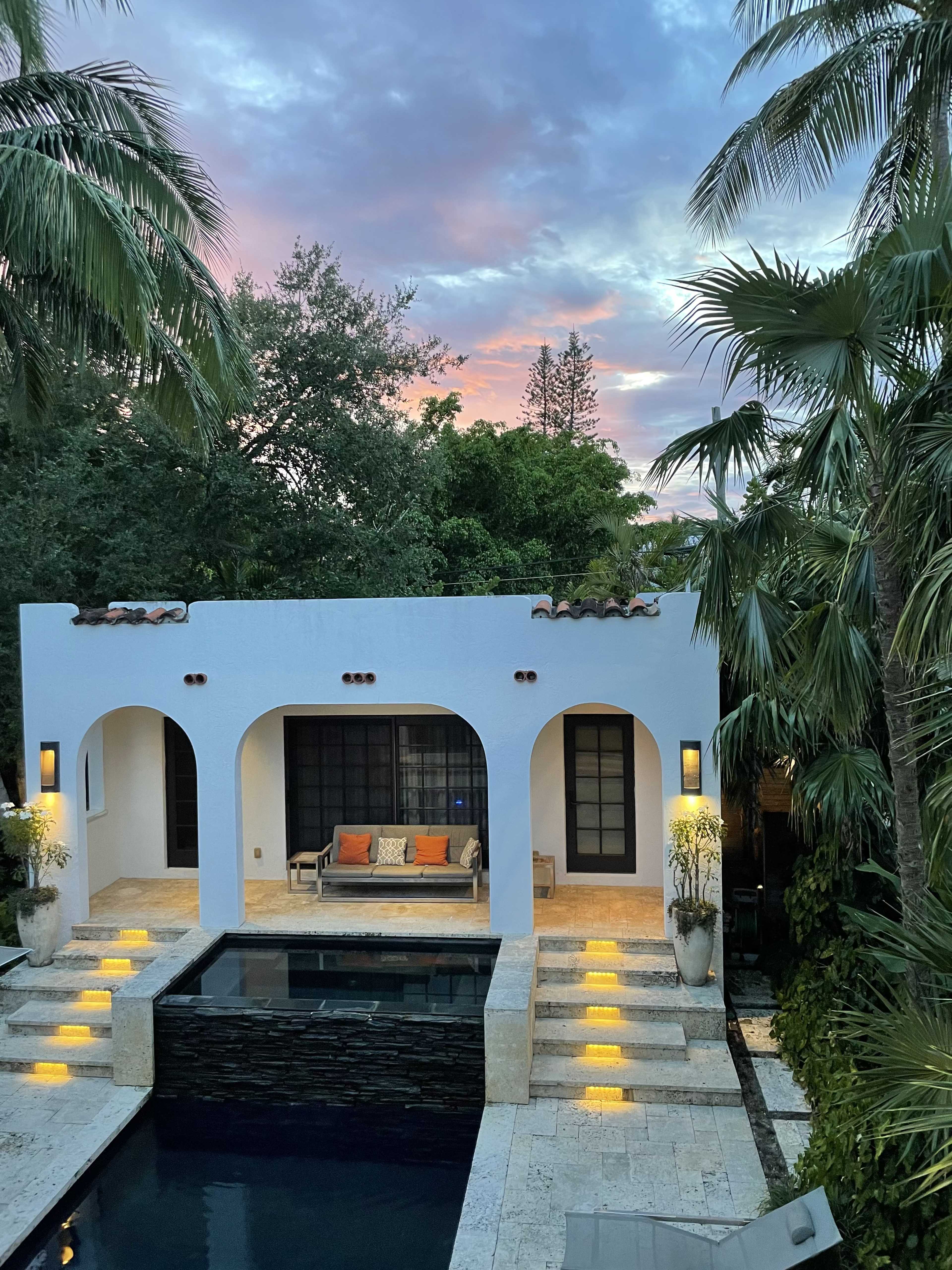 A modern white stucco house with arched doorways, a pool in front, and palm trees surrounding the property, all under a colorful sky at dusk.