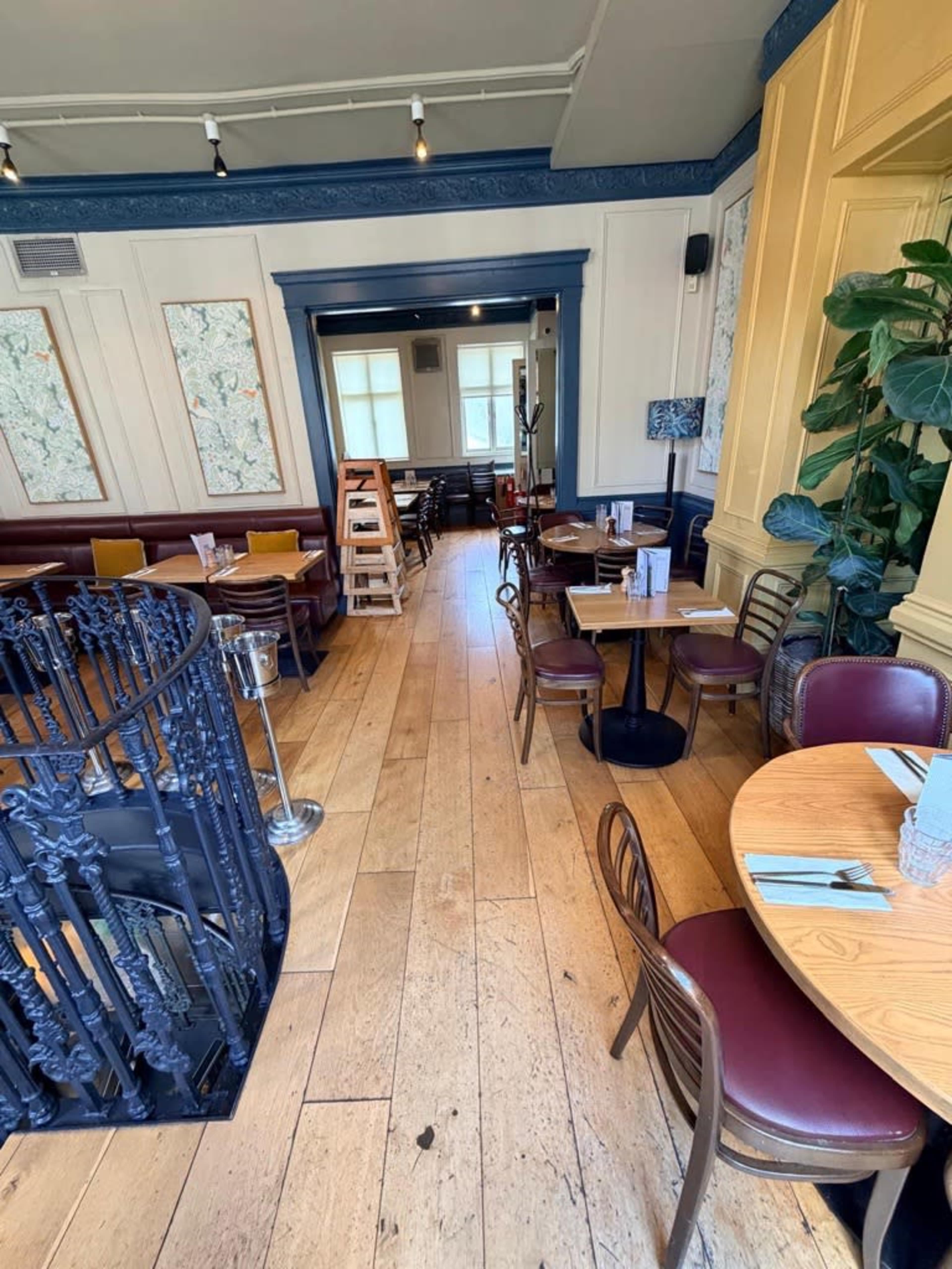 The image shows a restaurant interior with wooden flooring, circular and rectangular tables set for dining, and various seating arrangements along the walls.