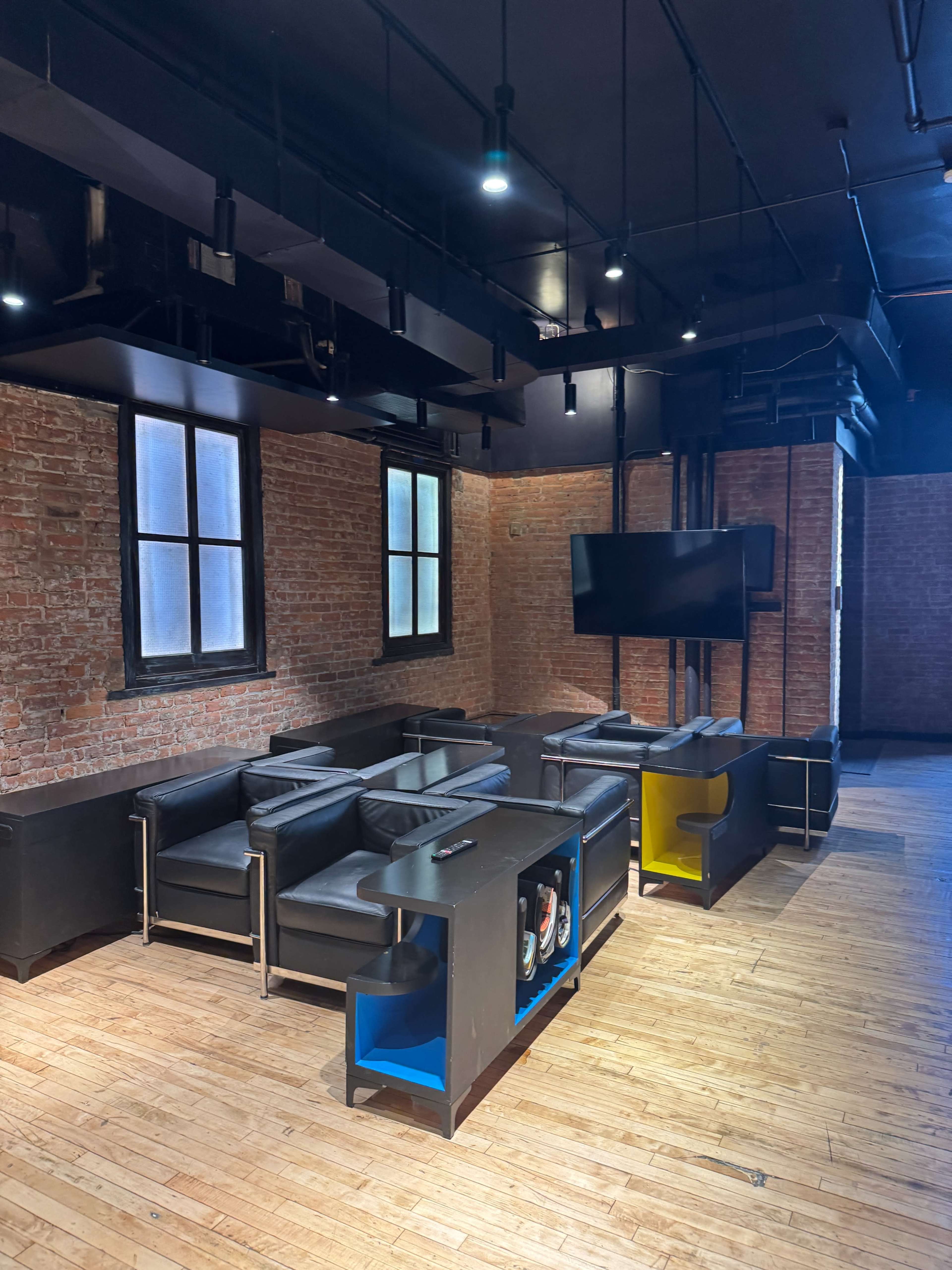 The image shows a spacious room with exposed brick walls, modern black furniture, and a television mounted on the wall.