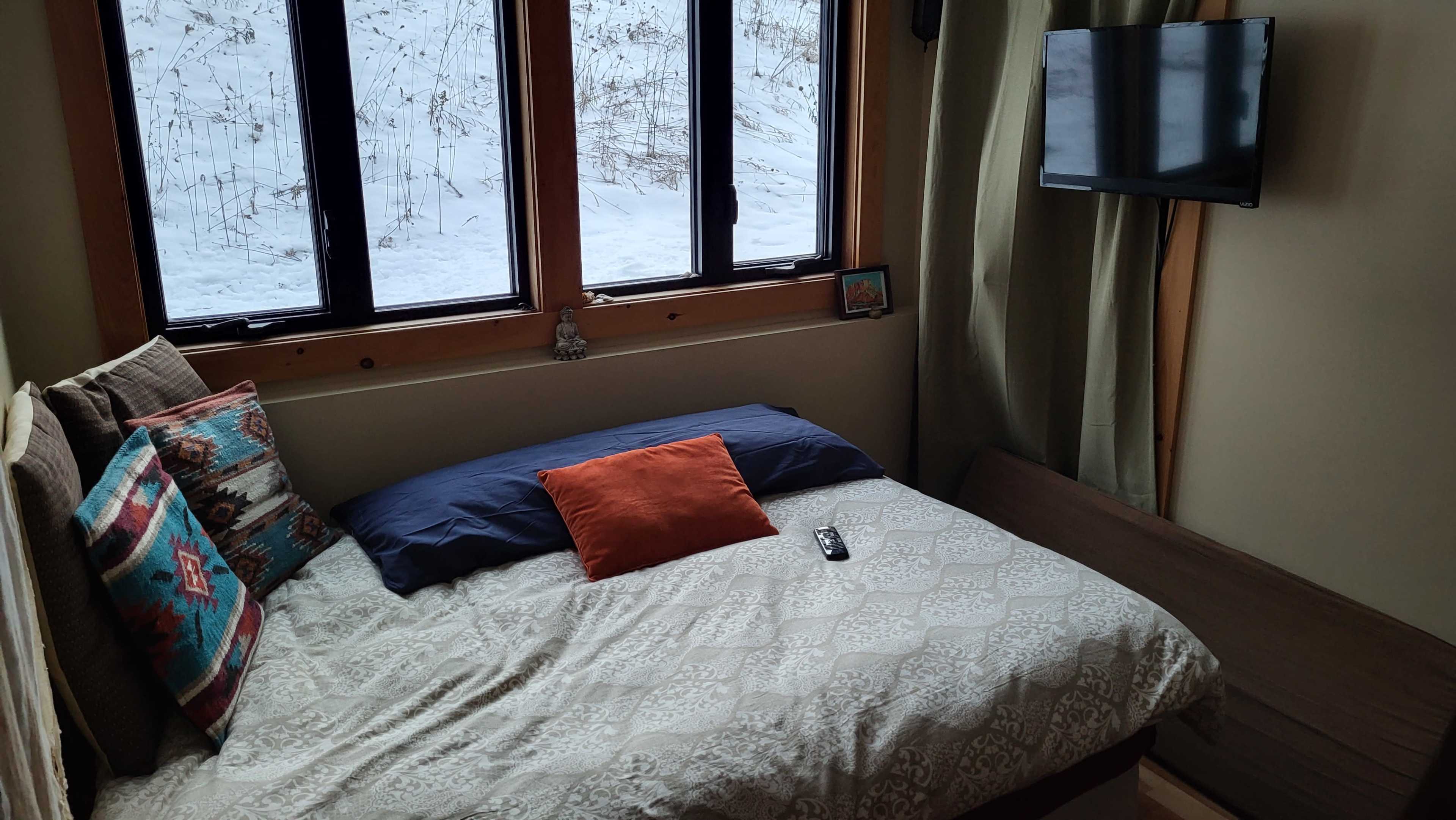 A cozy bedroom with a bed featuring patterned bedding, a few pillows, and a television mounted on the wall next to large windows overlooking a snowy landscape.