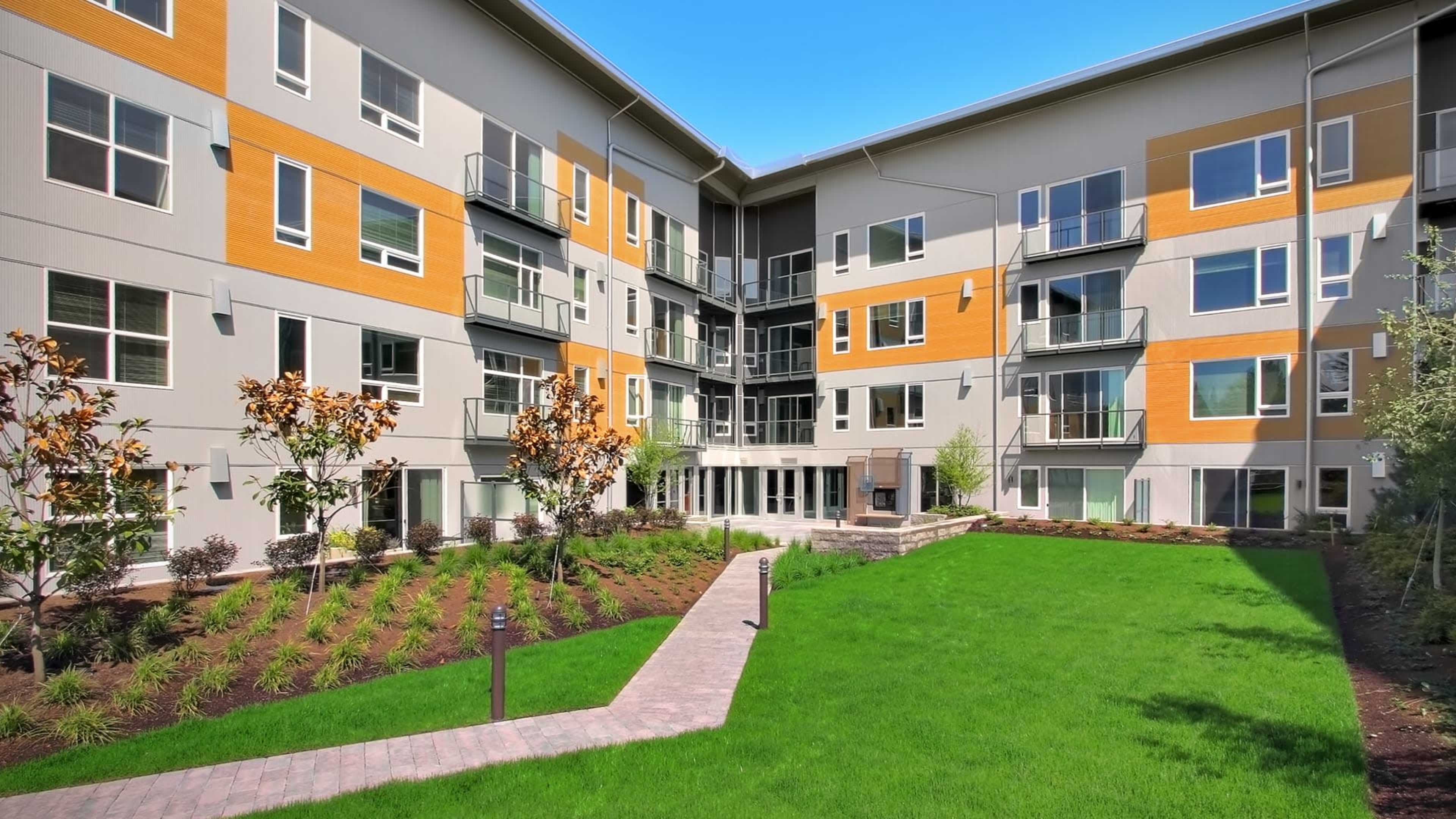 A modern apartment building with multiple floors surrounding a landscaped green area featuring a pathway and planted trees.