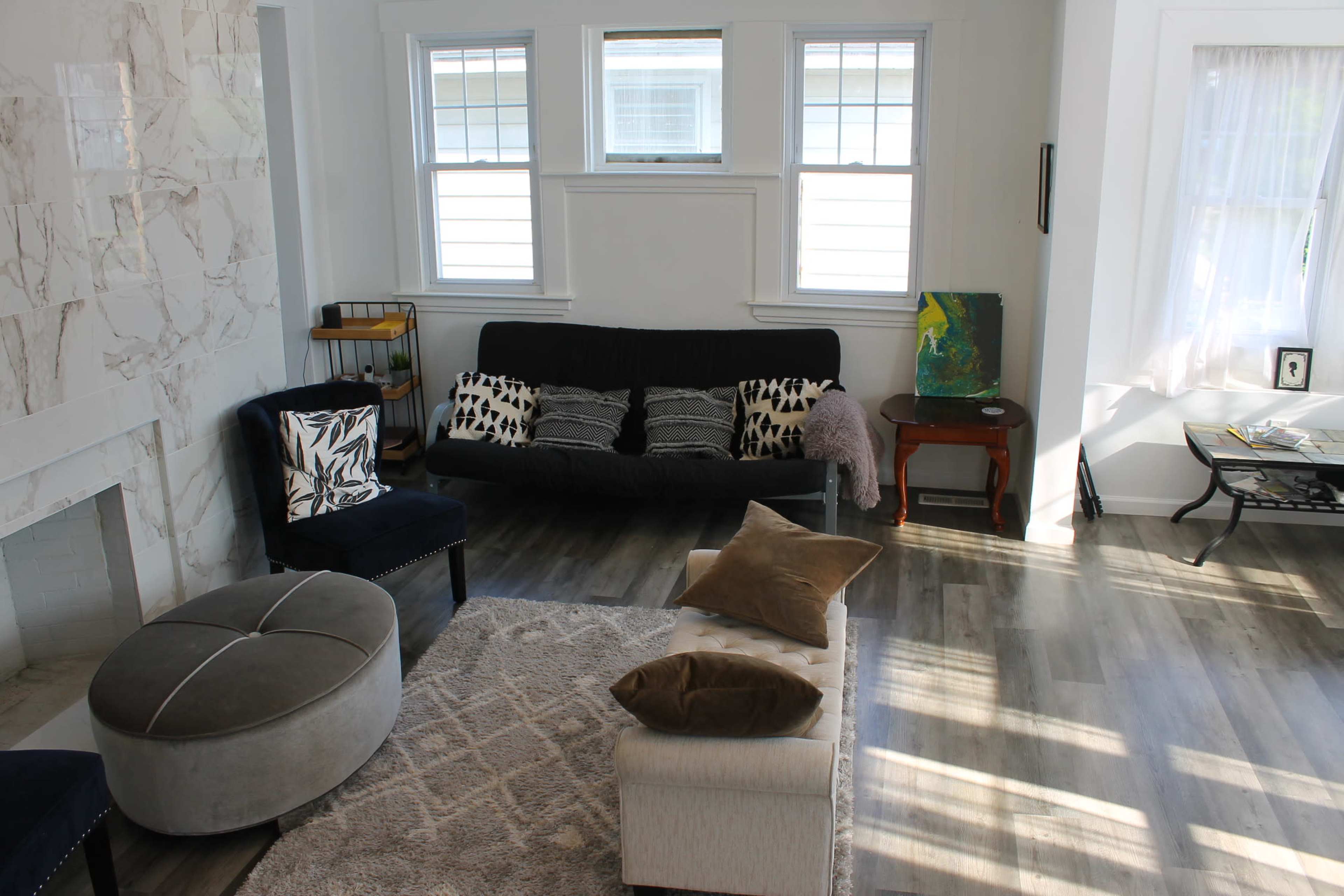 A living room with a sofa, armchairs, a round ottoman, and a coffee table, all arranged on a light-colored rug with wood flooring.