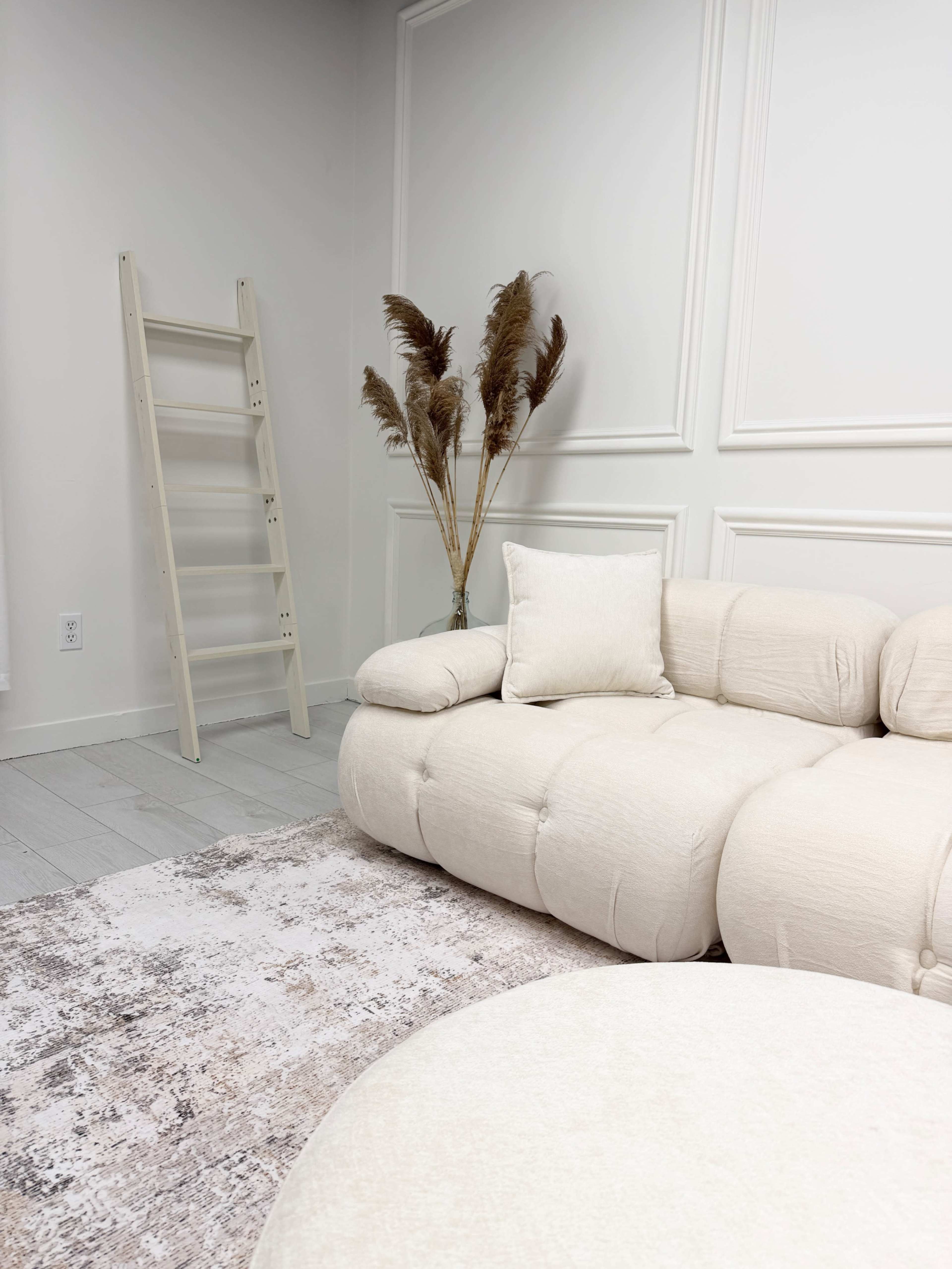 The image shows a light-colored living room featuring a curved sofa, a round ottoman, a decorative ladder, and a tall vase with dried pampas grass, all set against a softly textured wall.