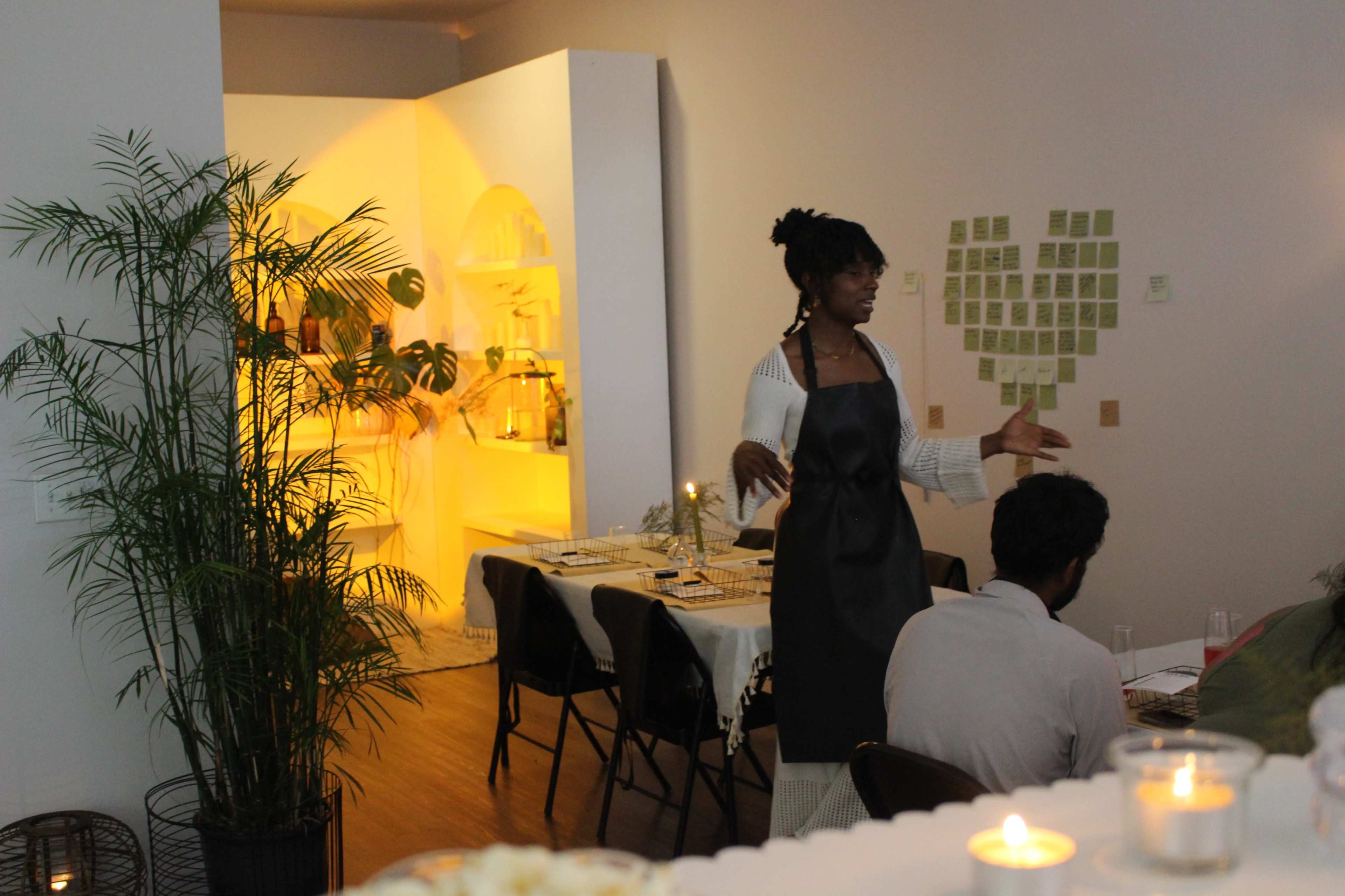A woman in an apron speaks to guests at a dining table in a softly lit room adorned with plants and decorative elements.
