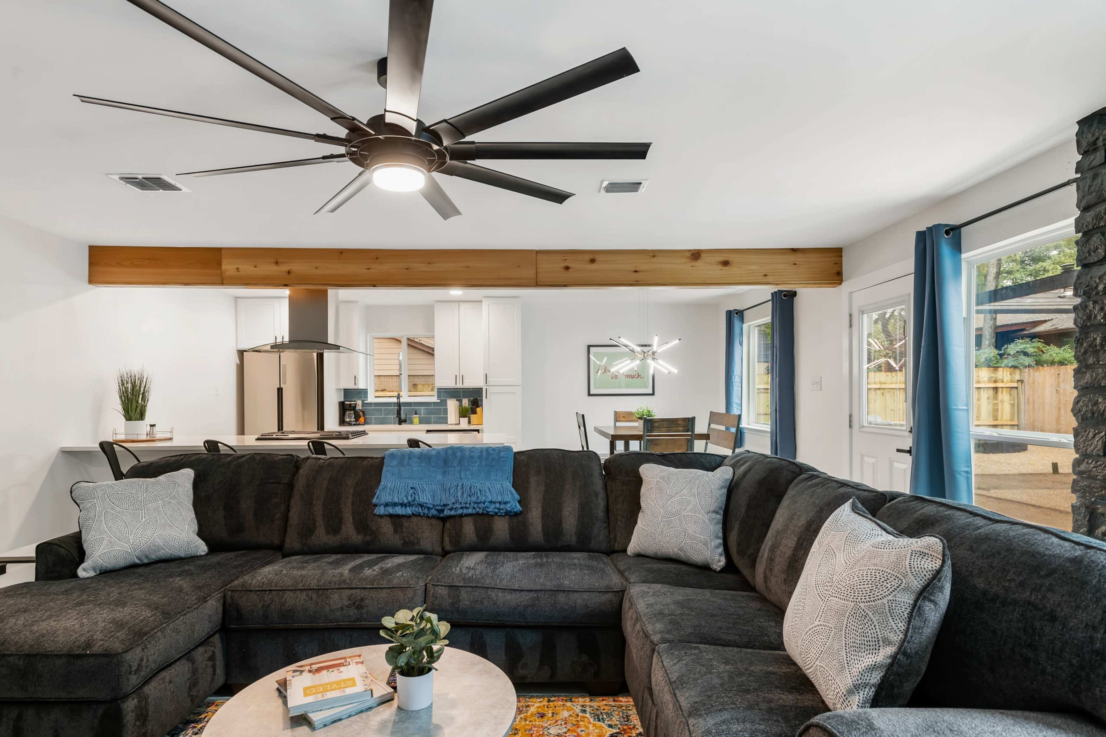 A spacious living room features a large sectional couch, a round coffee table, and a modern ceiling fan, with an open layout leading to a kitchen area.