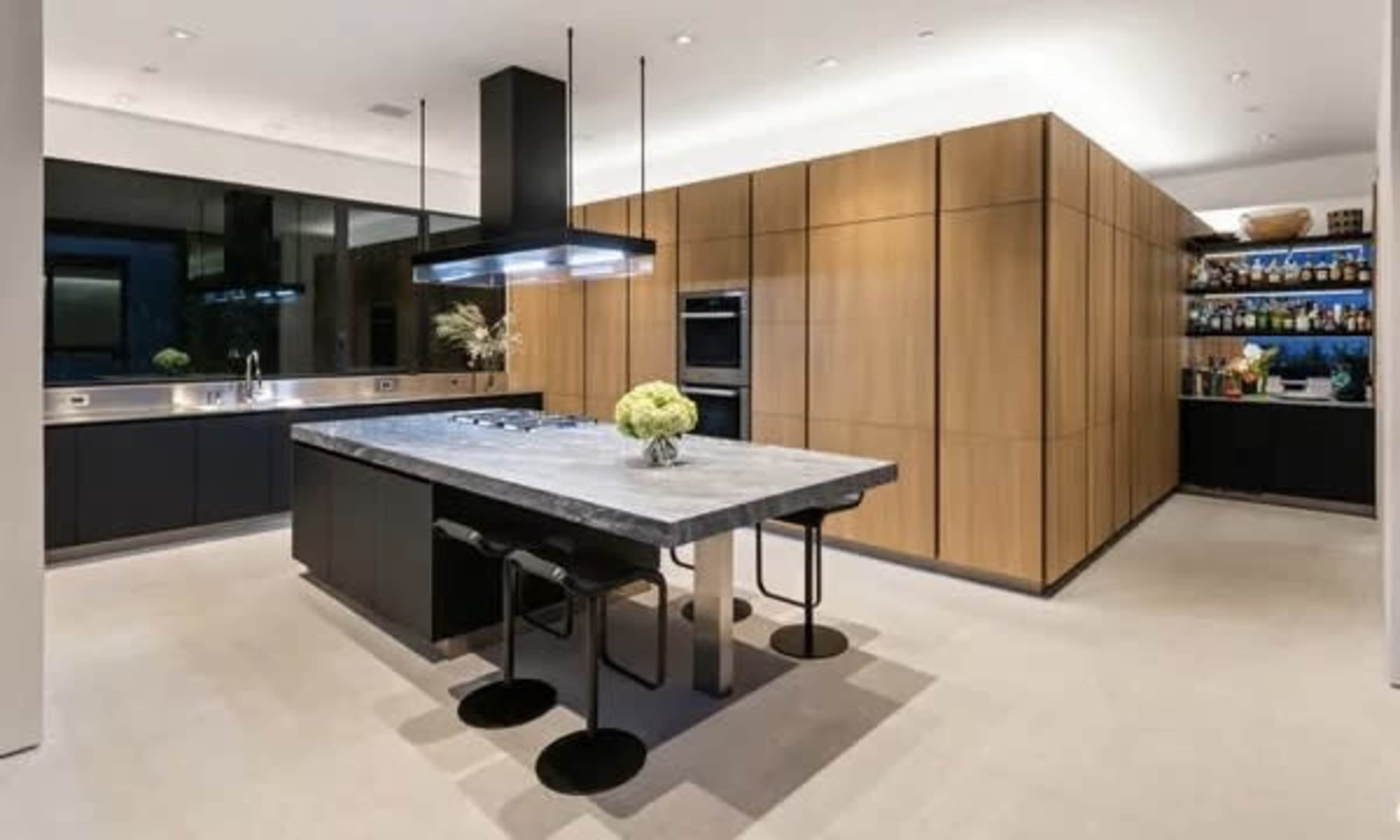 The kitchen features a central gray island with four black stools, surrounded by wooden cabinetry and modern appliances.