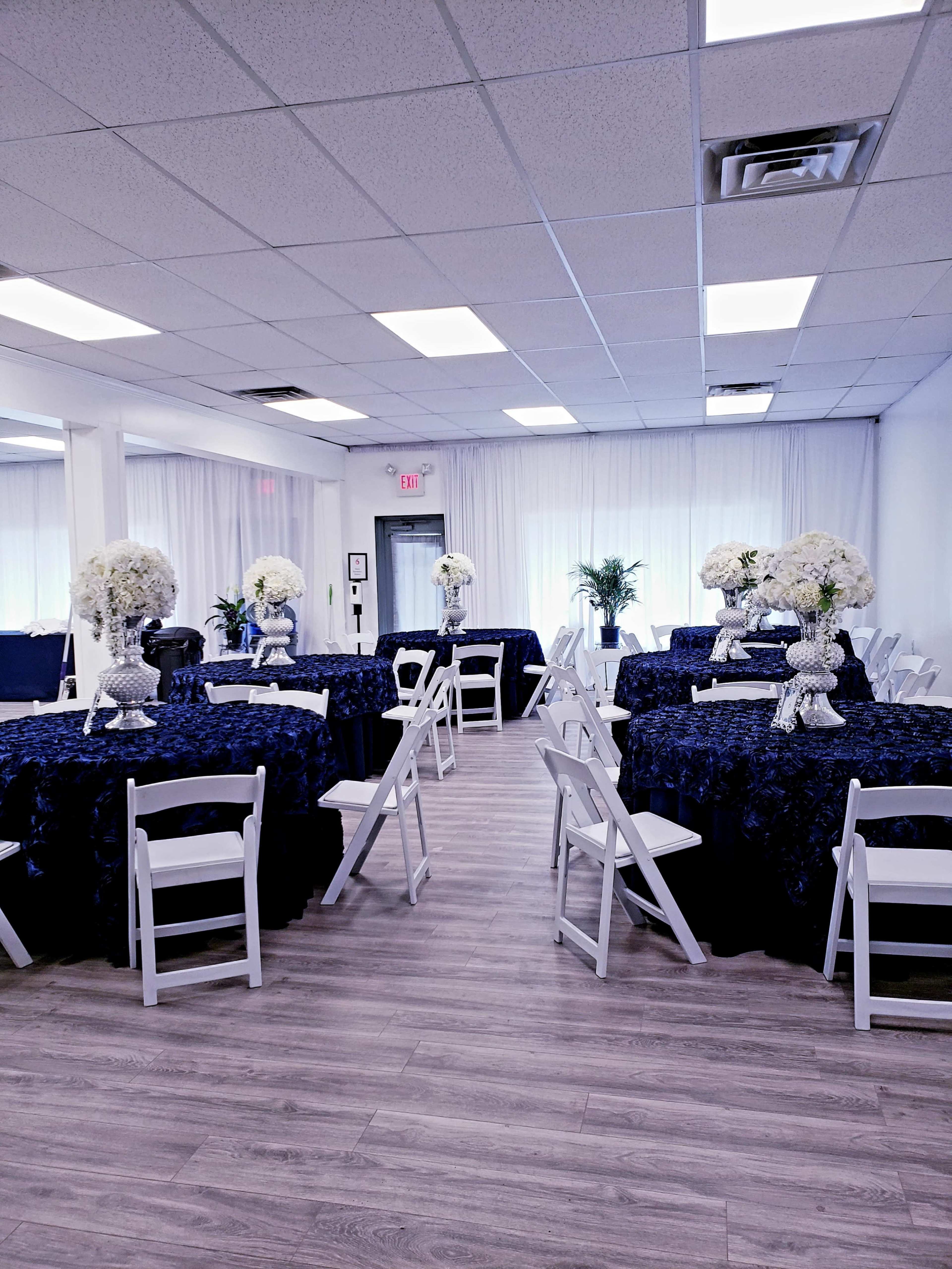The scene shows a banquet hall set up with round tables covered in dark blue tablecloths, adorned with floral centerpieces, and surrounded by white folding chairs.