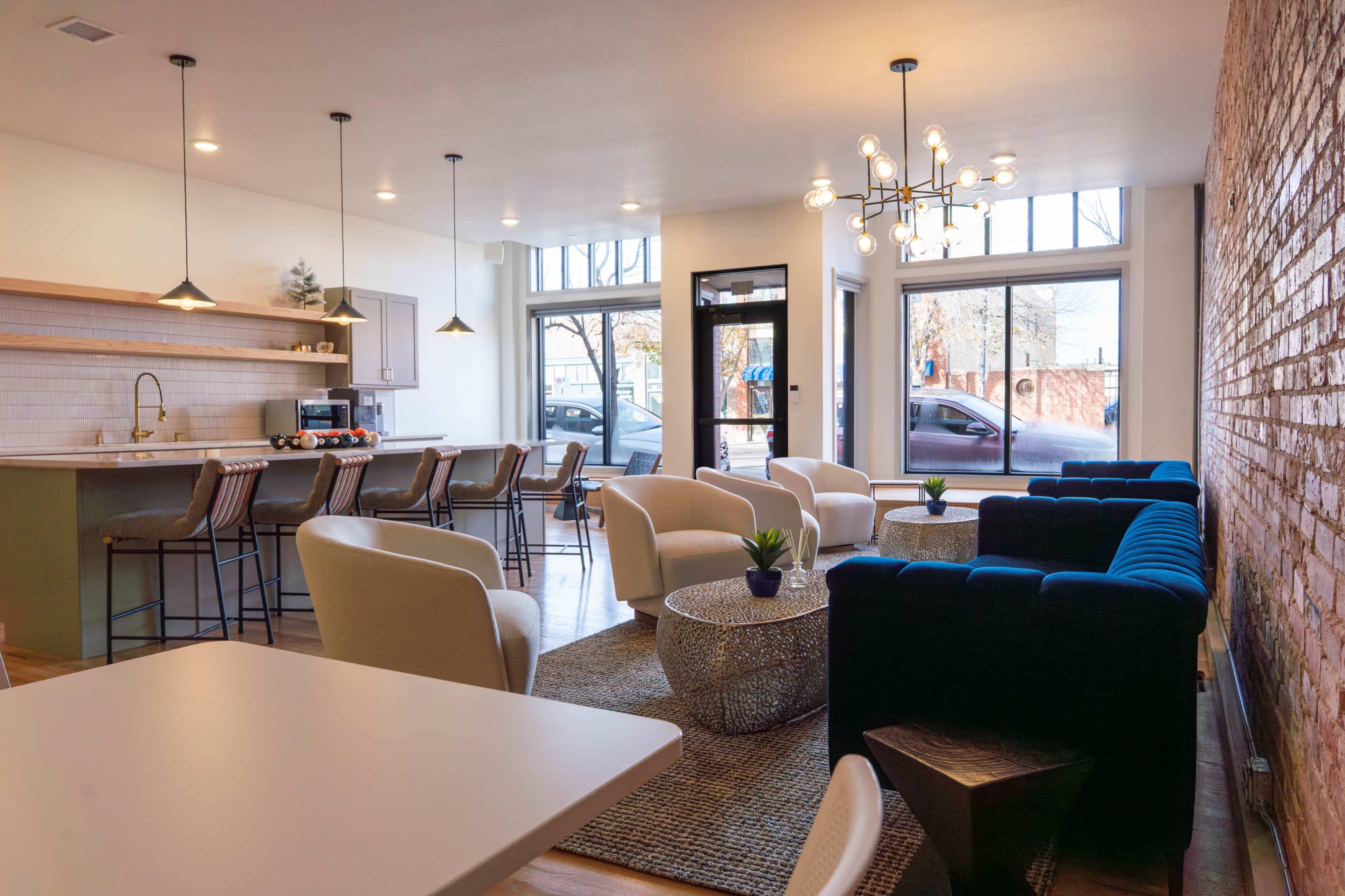 The image shows a modern, open lounge area featuring a kitchen space, seating arrangements with armchairs and a couch, a coffee table, and large windows allowing natural light.