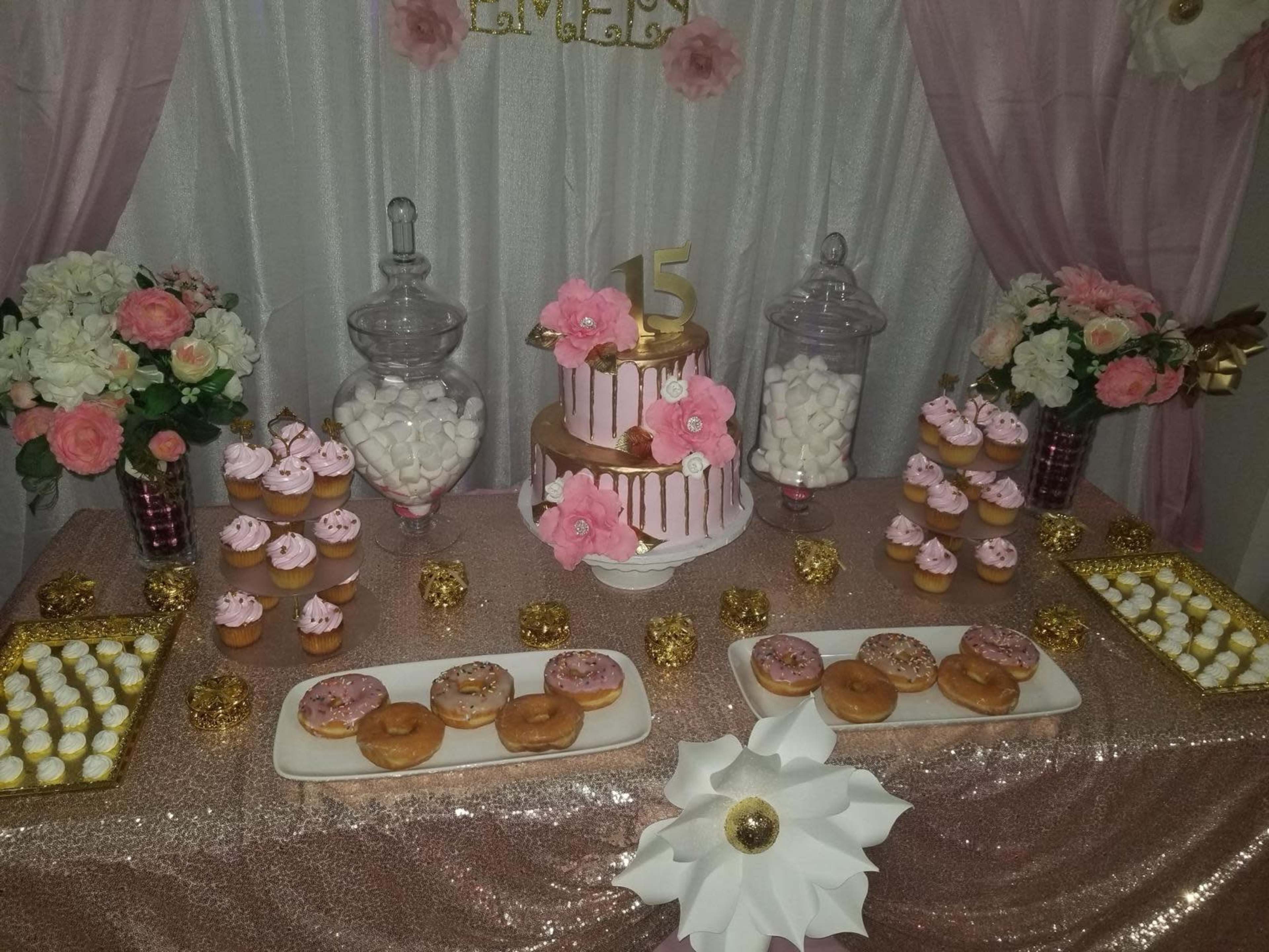 A decorated dessert table featuring a three-tiered cake adorned with flowers, alongside various pastries, including cupcakes and donuts, arranged on plates and in jars.