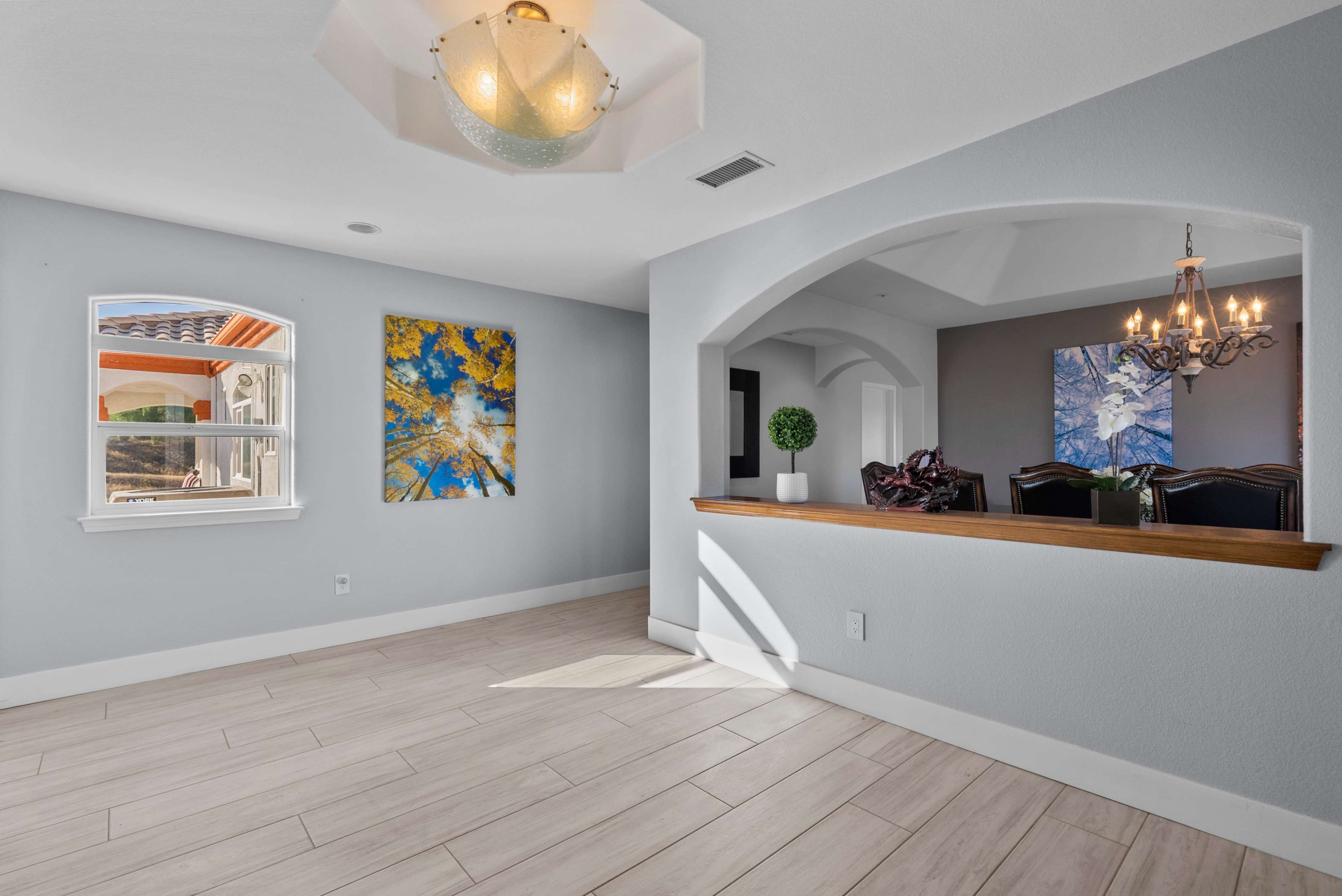 A spacious interior room features large windows, light-colored walls, and decorative paintings, with a view of a dining area beyond an arched passage.