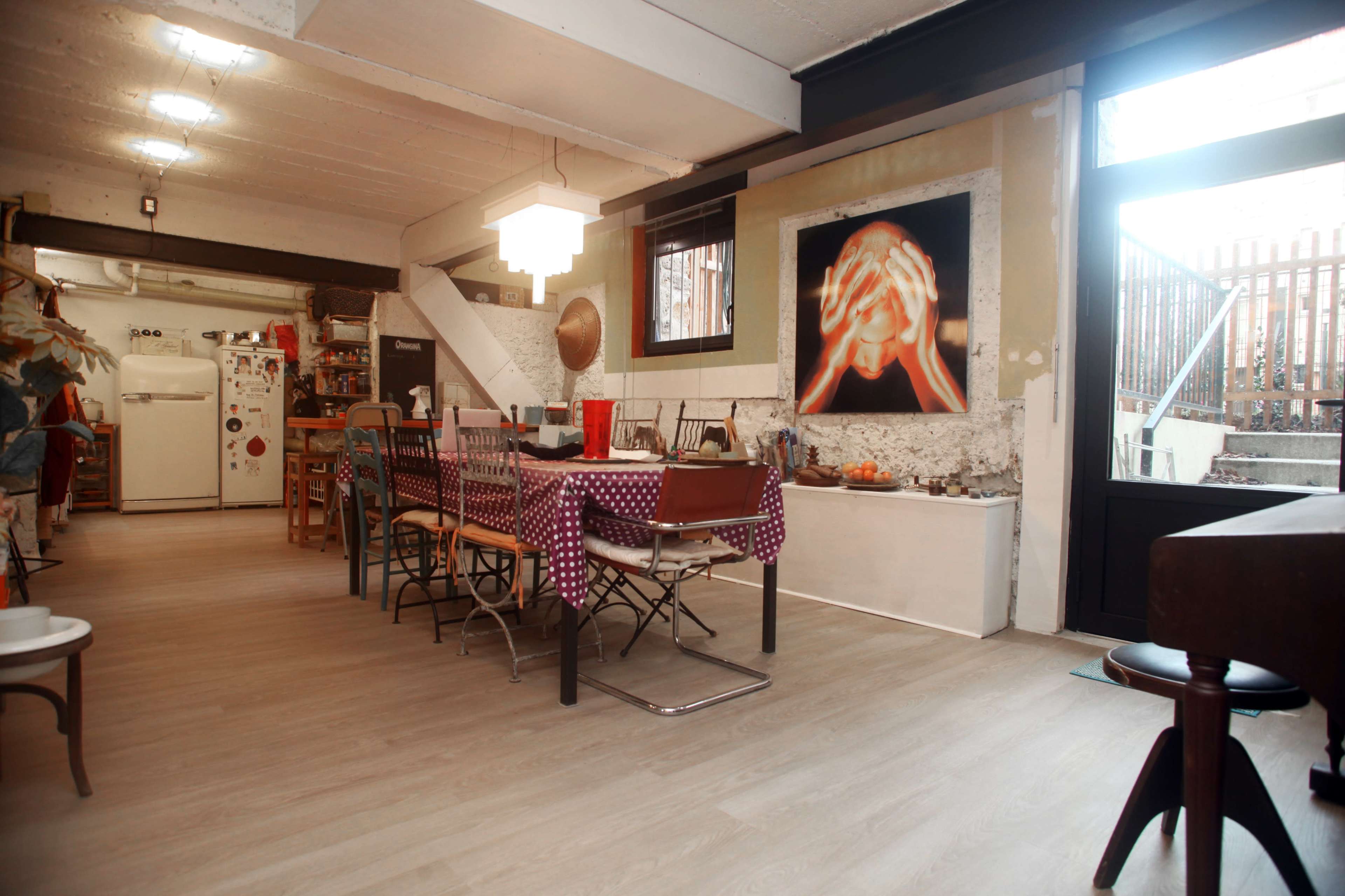 The image shows a spacious, well-lit room featuring a dining table with chairs, a kitchenette, and an artwork on the wall.