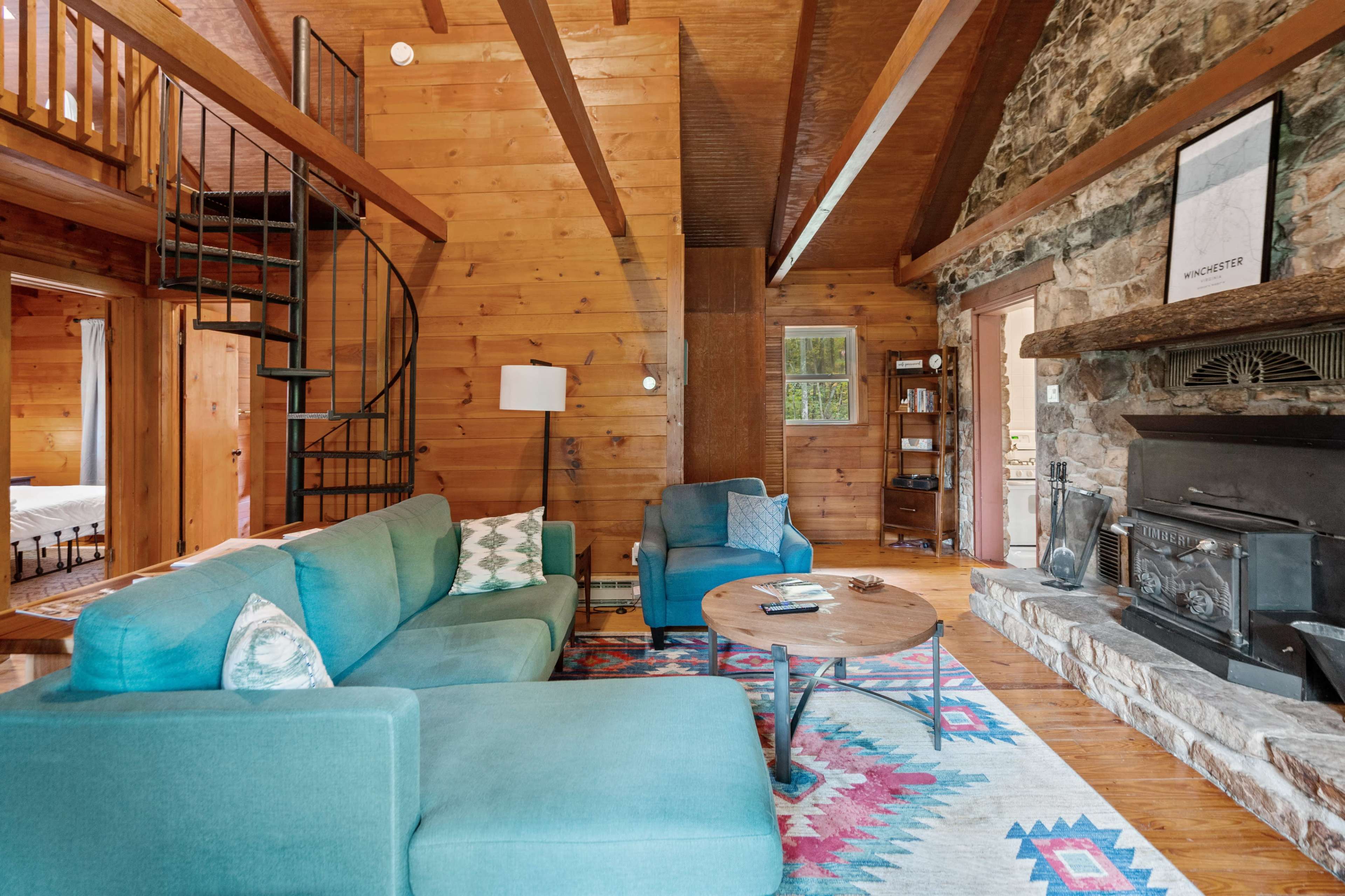 The interior of a rustic cabin features a living area with a teal sectional sofa, a round wooden coffee table, a stone fireplace, and a spiral staircase leading to an upper level.