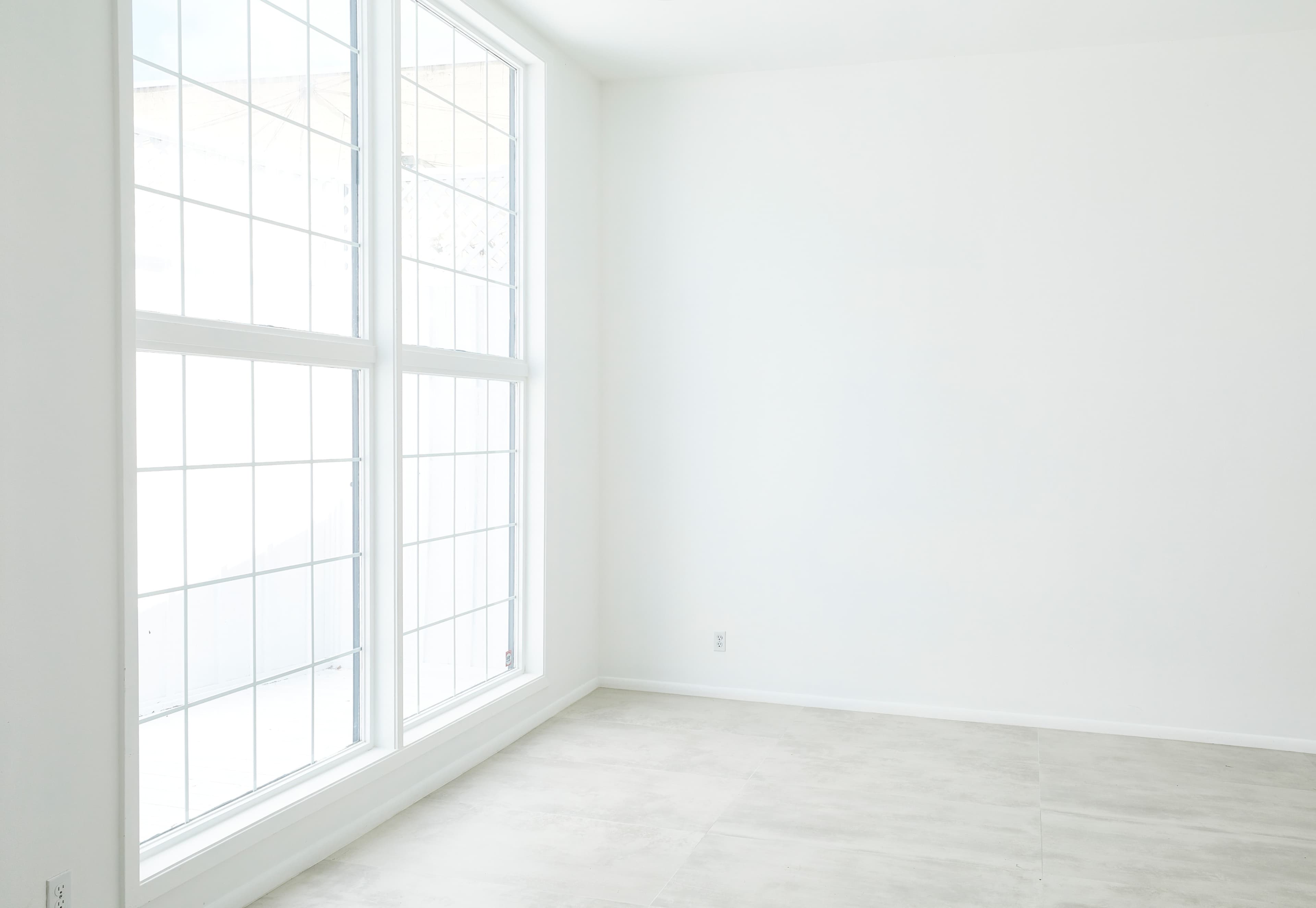 A spacious room features two large windows with grid designs letting in natural light, and has plain white walls and light gray flooring.