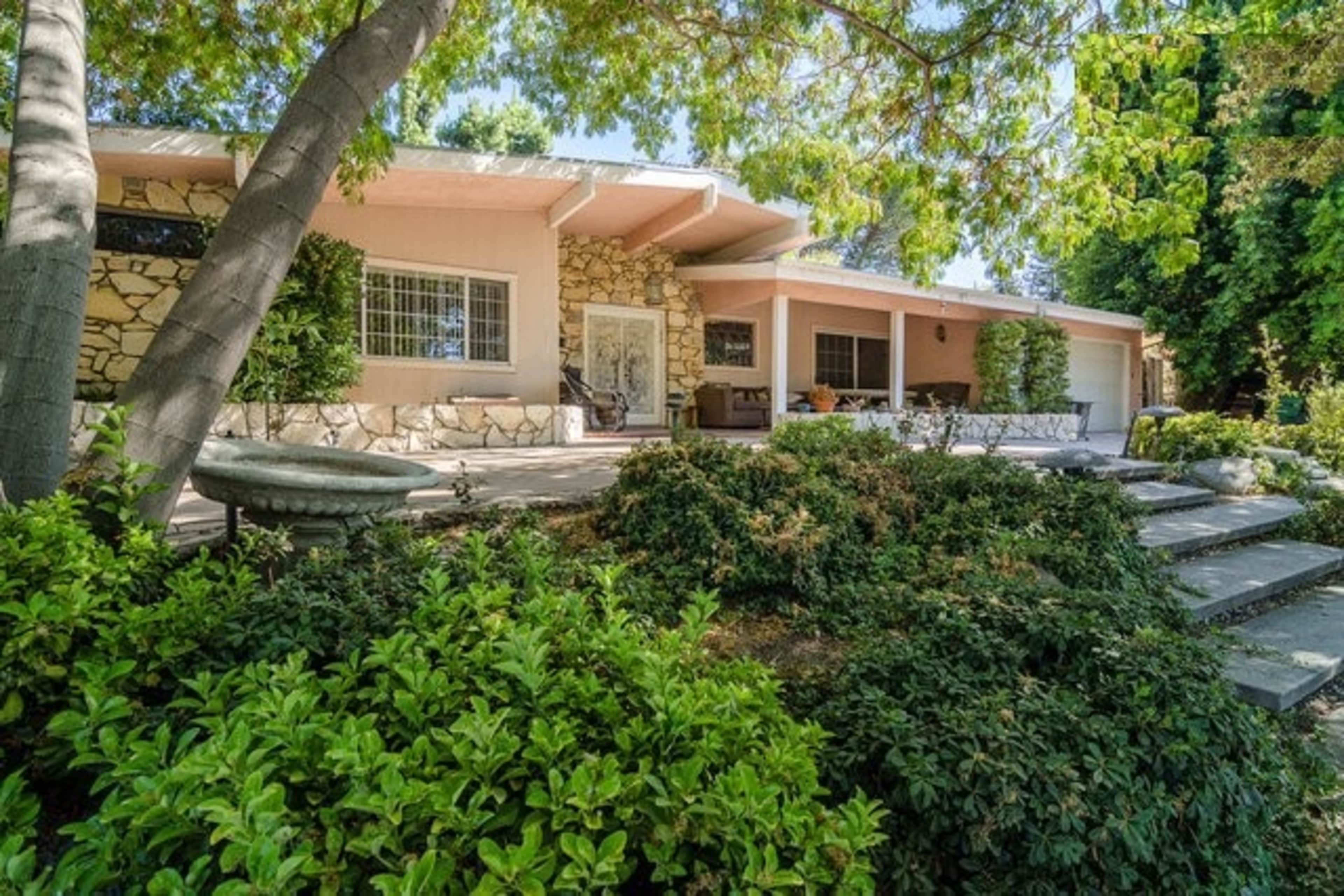 A single-story house with a stone façade, surrounded by landscaped greenery and a stone walkway.