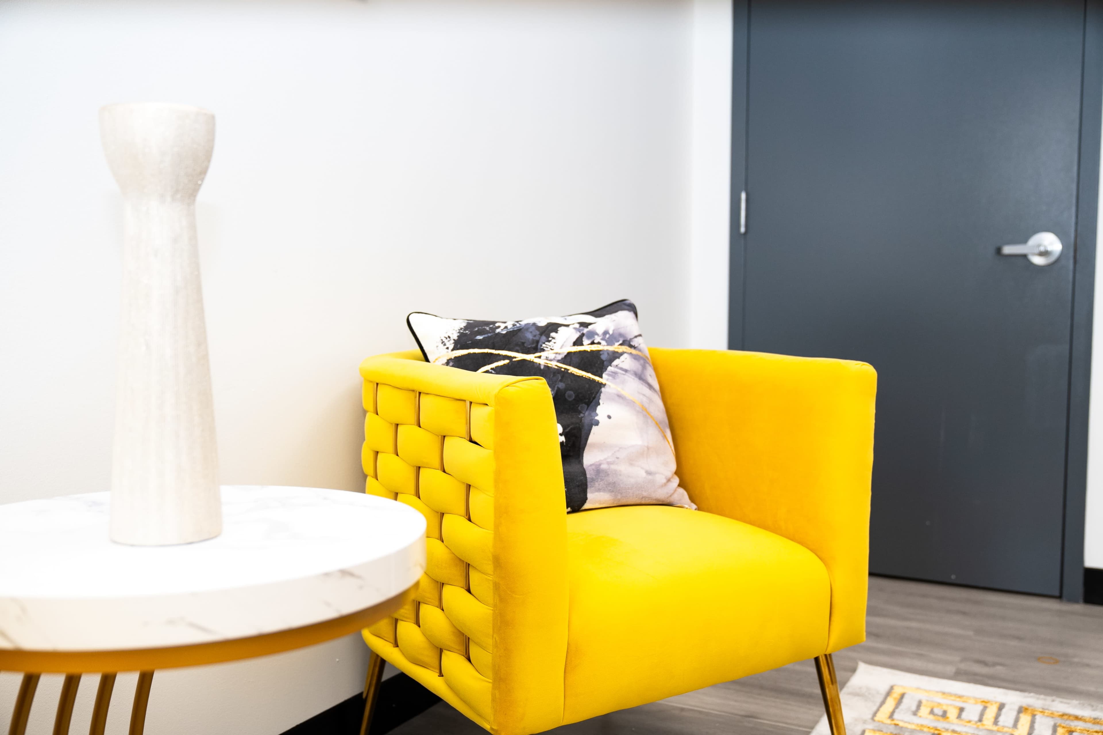 A yellow armchair with tufted sides is positioned next to a round marble-top table and a dark door.