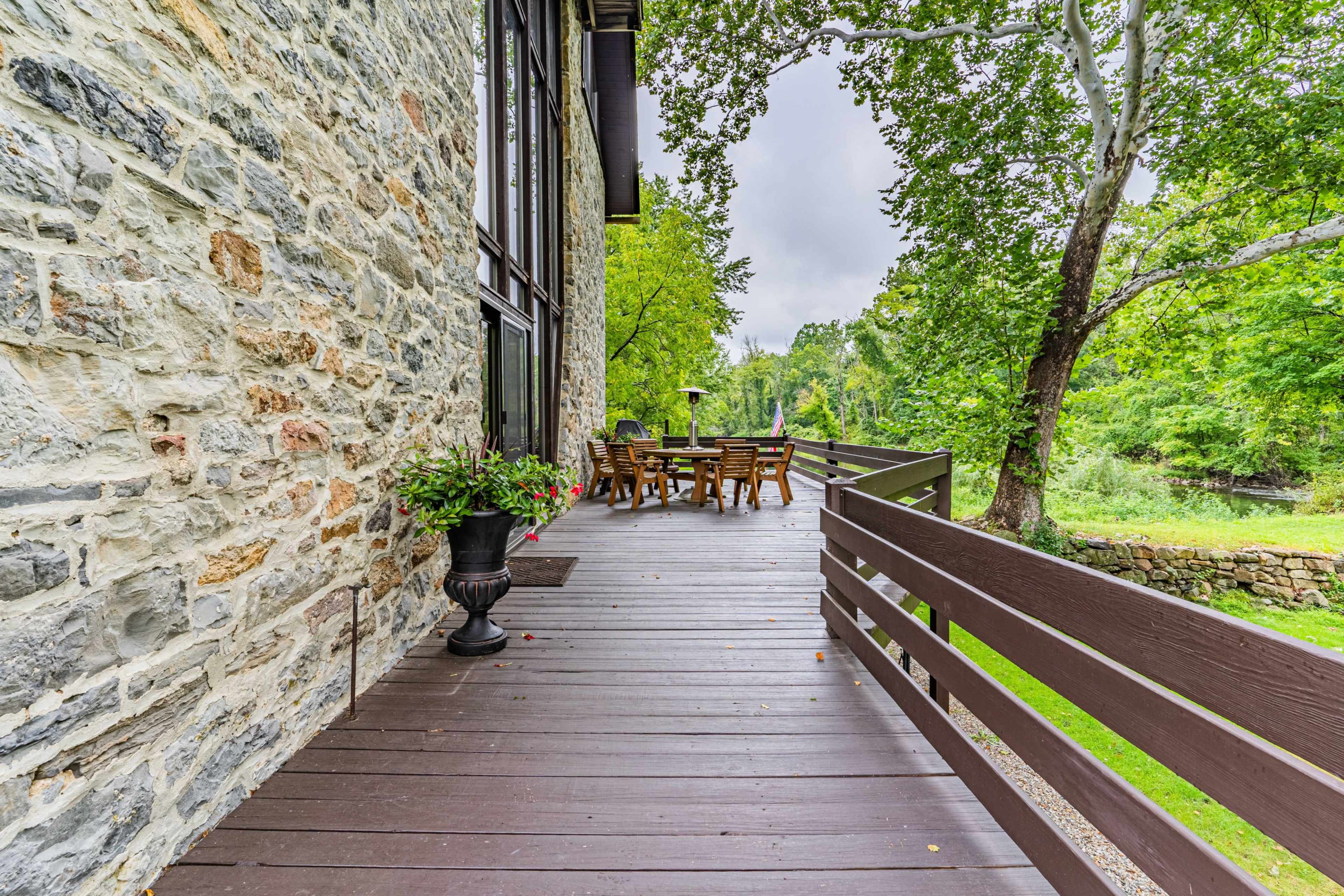 A wooden deck extends alongside a stone house, leading to a set of outdoor dining furniture under the shade of trees.