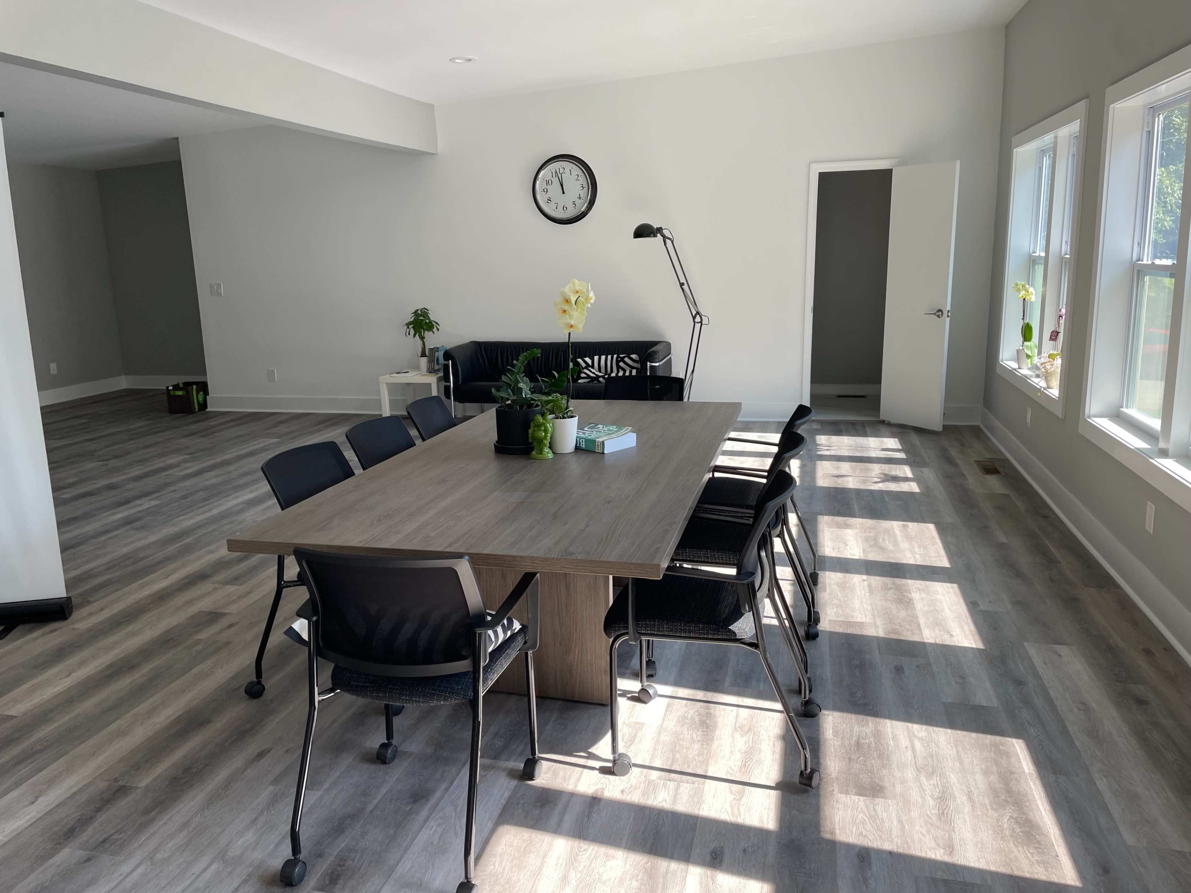 A modern meeting room features a large wooden table surrounded by black chairs, with large windows allowing natural light to fill the space.