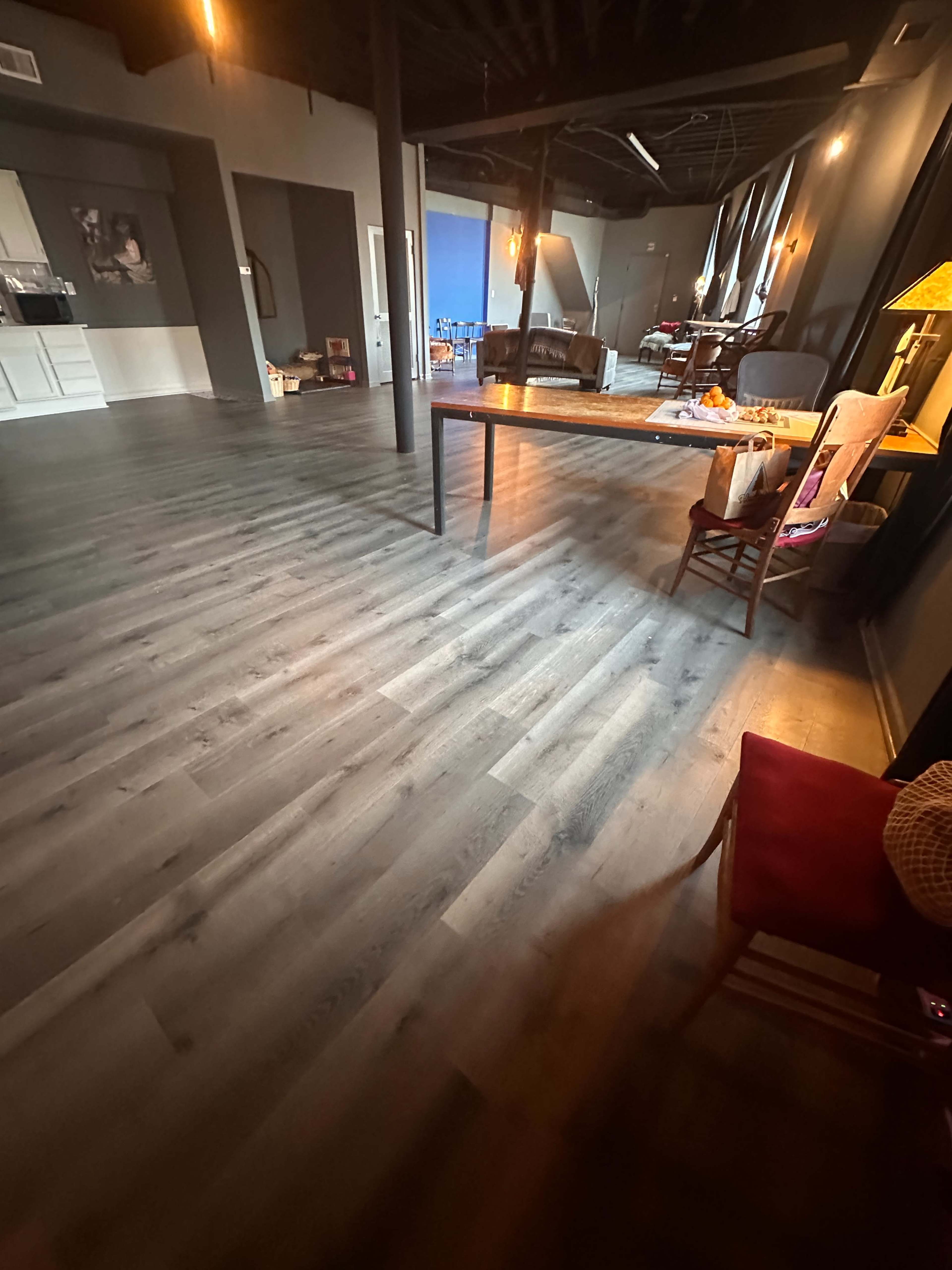The image shows a spacious, modern interior with gray laminate flooring, featuring a long table, chairs, and ambient lighting.
