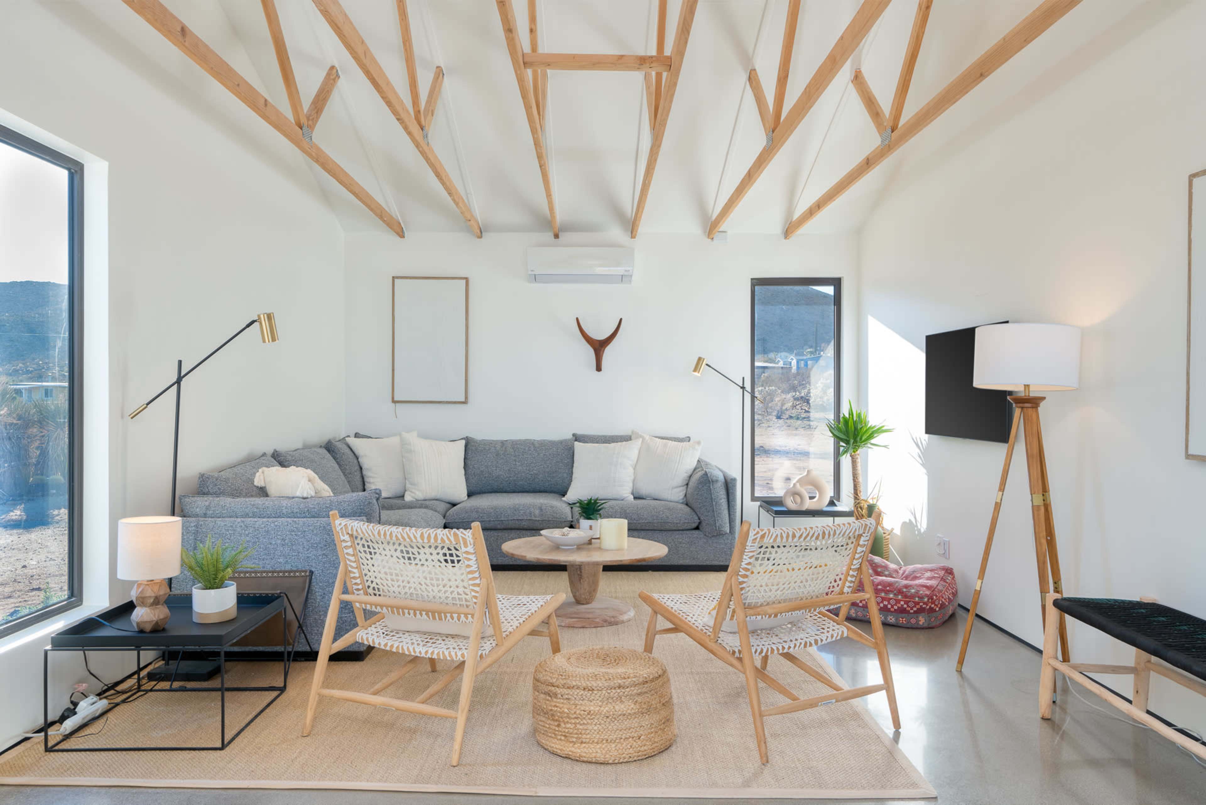 A modern living room features a gray sectional sofa, two wooden chairs, and a round coffee table, with large windows and minimal decor.