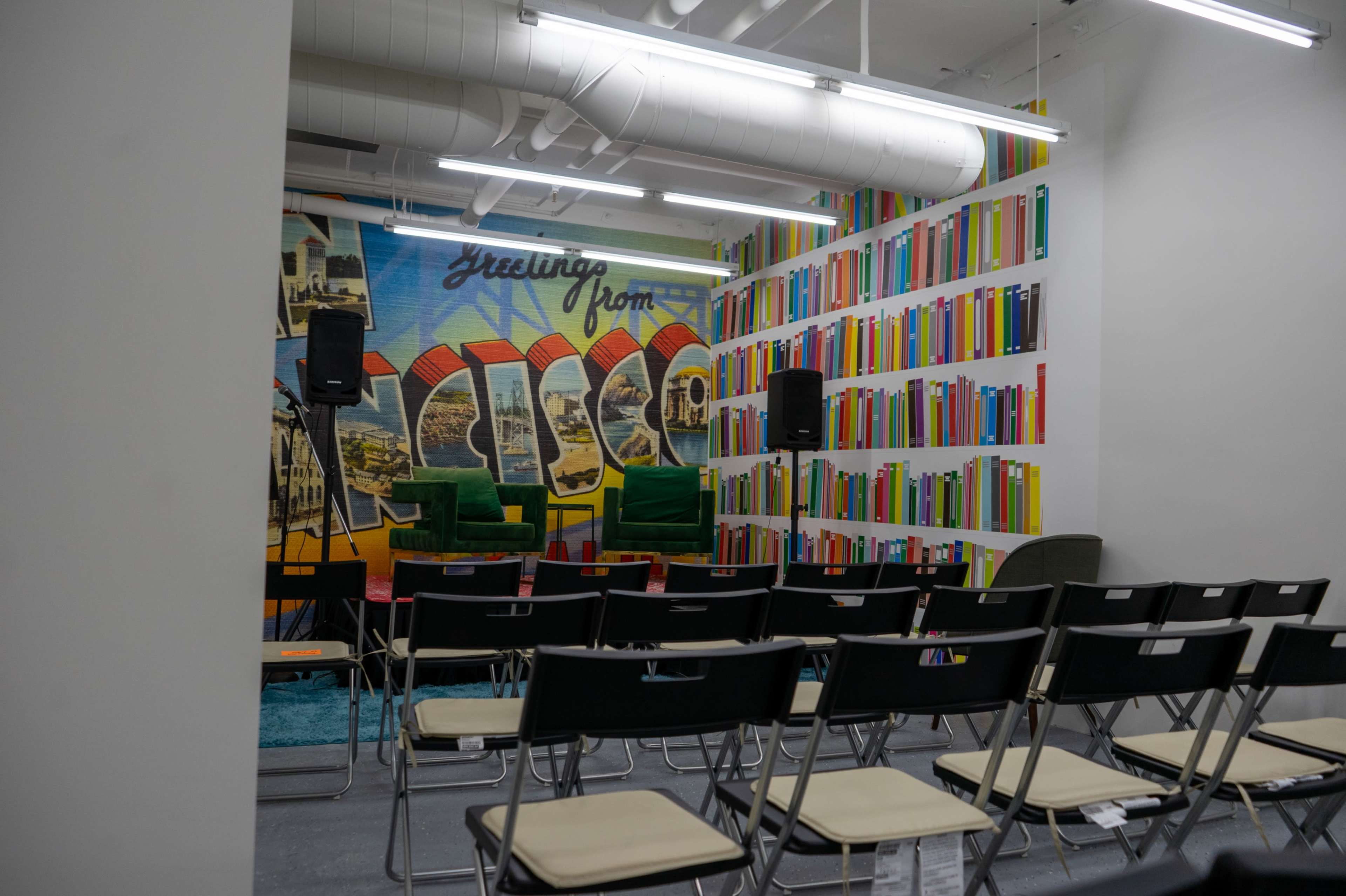 The image shows a small event space with two green chairs and a colorful mural on the back wall, surrounded by rows of empty folding chairs.
