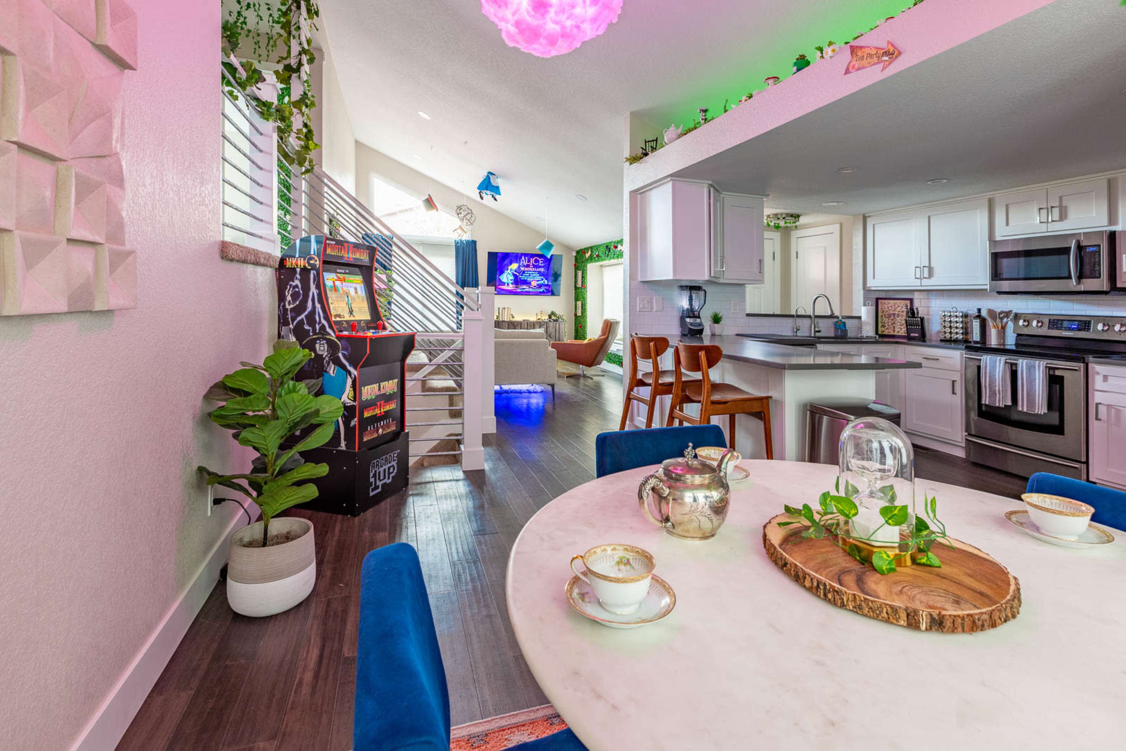 A bright, modern kitchen and dining area features a marble table with tea cups, an arcade game, and a view of a living room decorated with plants and colorful lighting.