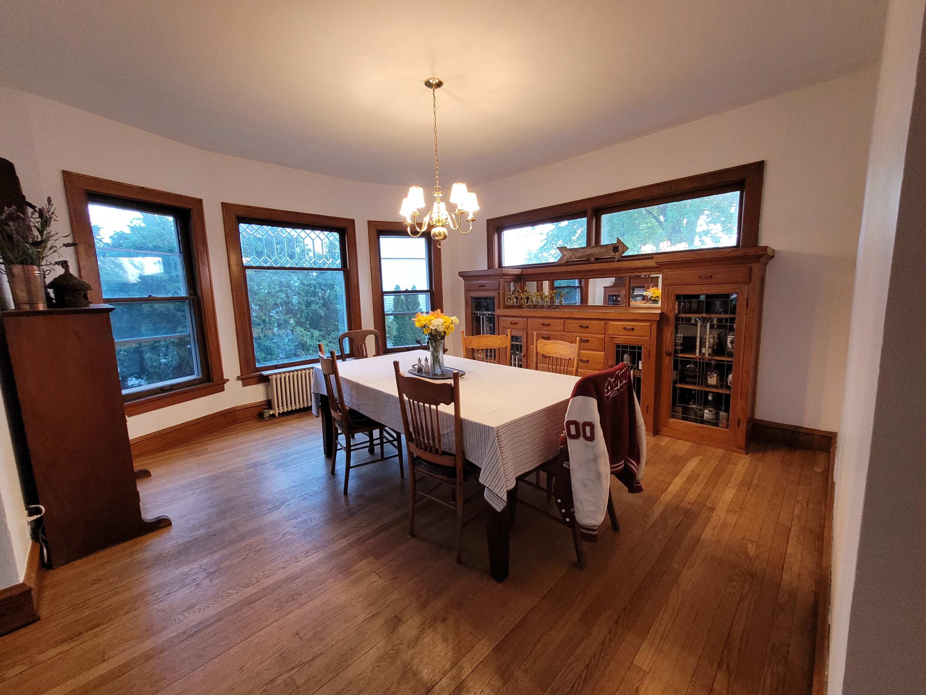 A spacious dining room features a large table surrounded by chairs, with a china cabinet against the wall and windows letting in natural light.