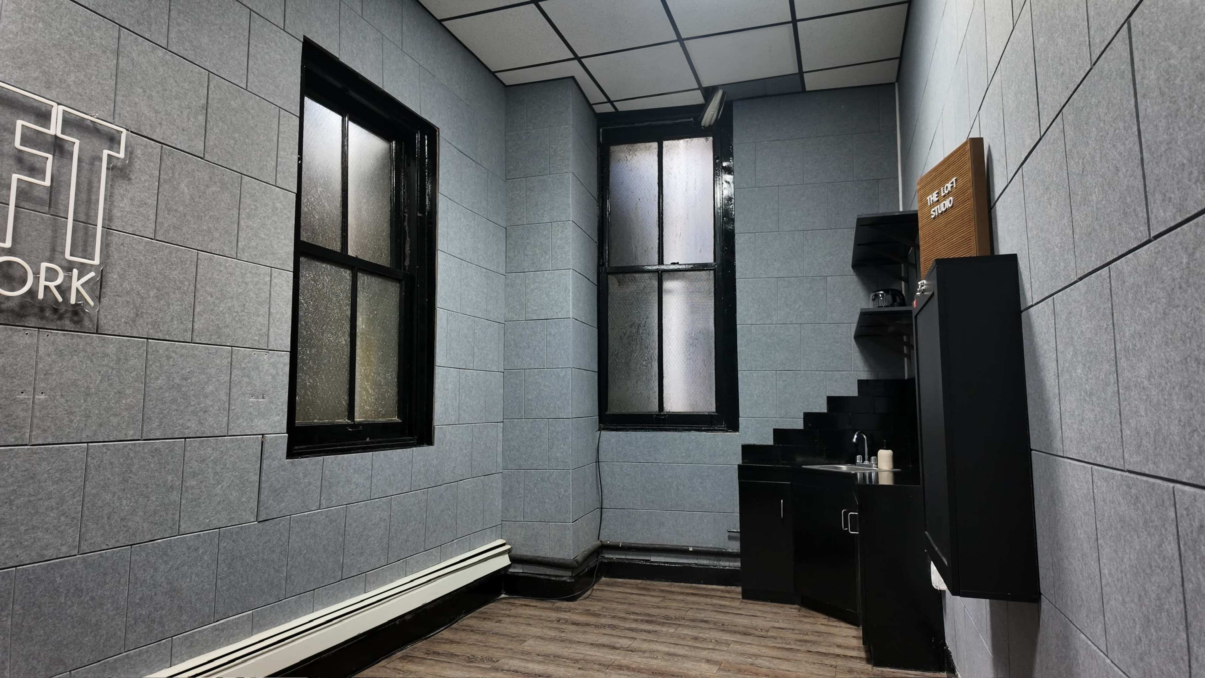 The image shows a small, gray-walled room with two windows, a sink, and shelves in one corner.