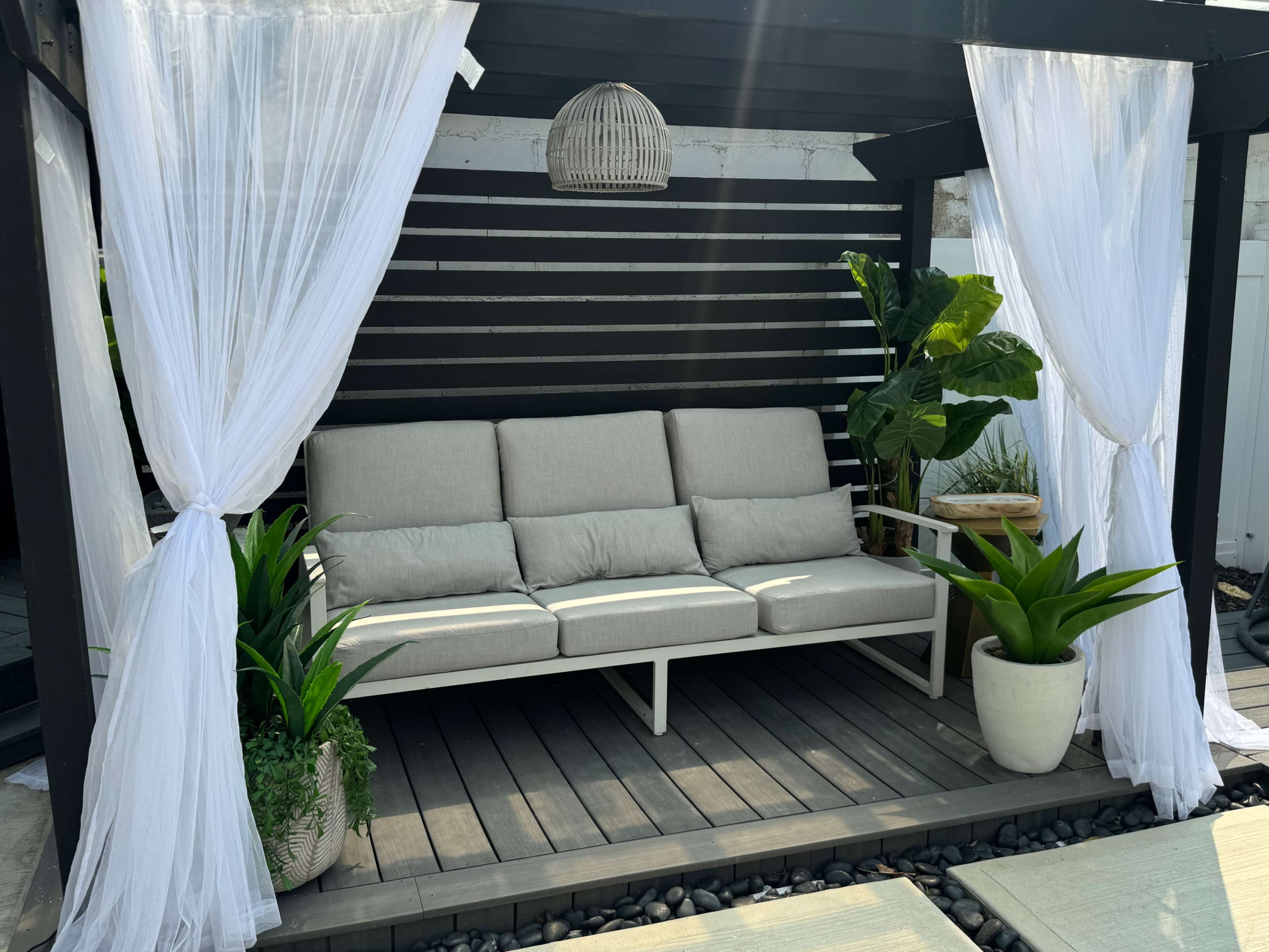 The image shows a cozy outdoor seating area with a light-colored sofa, potted plants, and white curtains draping from a black pergola.