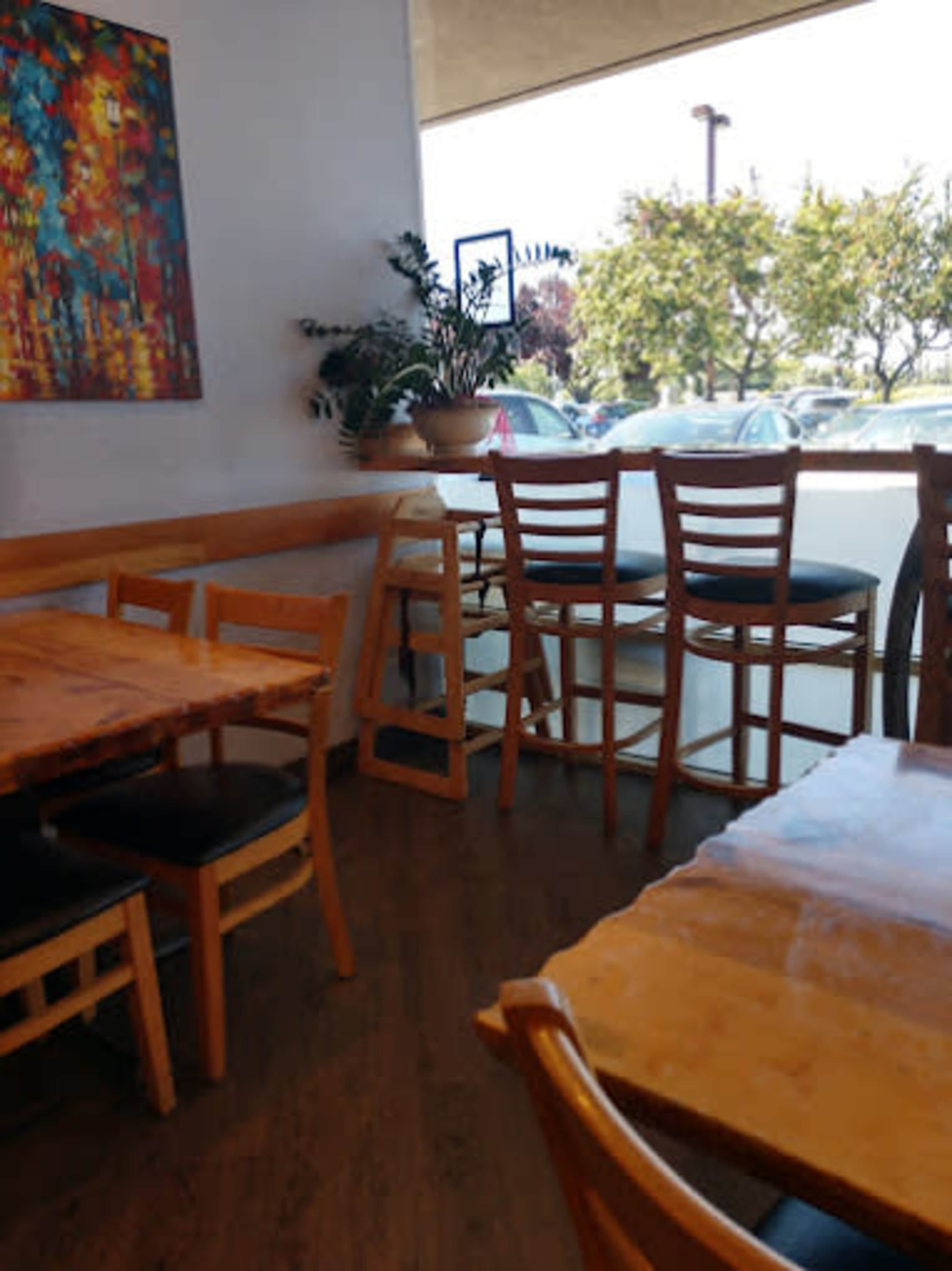 The image shows the interior of a café with wooden tables and chairs, a large window letting in natural light, and plants placed on a ledge.