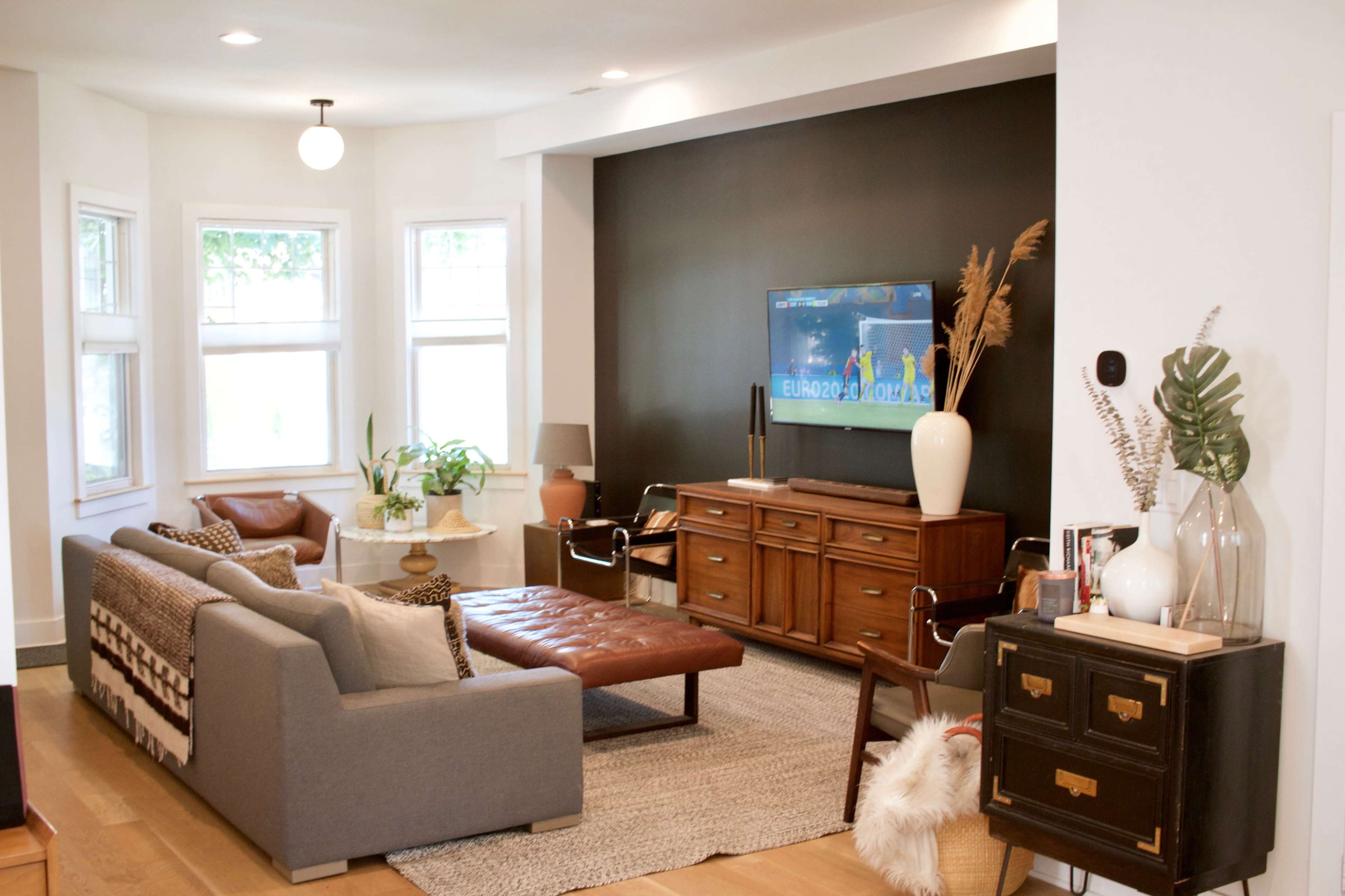 The living room features a gray sectional sofa, a leather coffee table, and a dark accent wall with a television, surrounded by large windows and decorative plants.