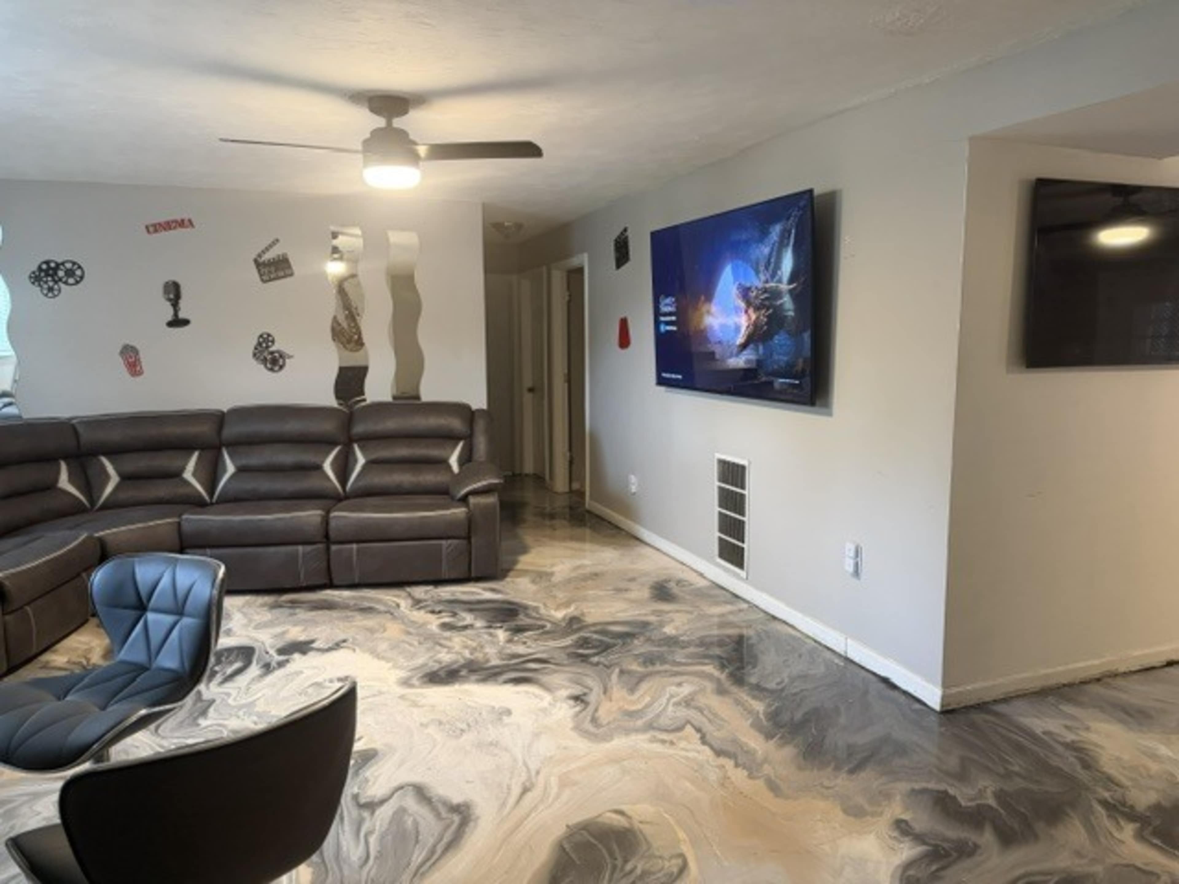 The image shows a modern living room with a large sectional sofa, two wall-mounted TVs, and a unique marbled floor design.